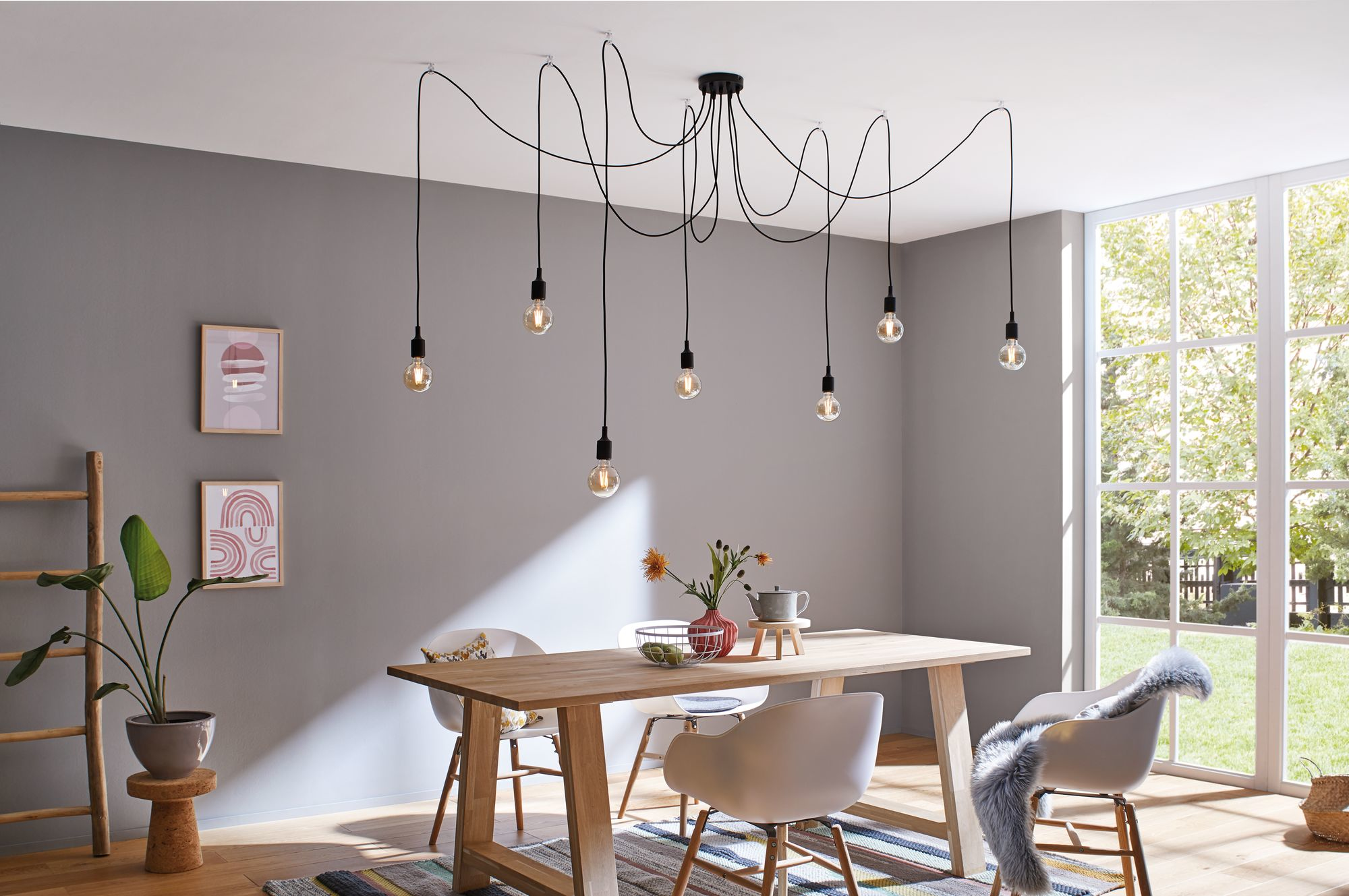 Jadalnia: drewniany stół, białe krzesła, wiszące lampy, okno z drzewami. Grafika na ścianie, roślina na stołku i dywan.