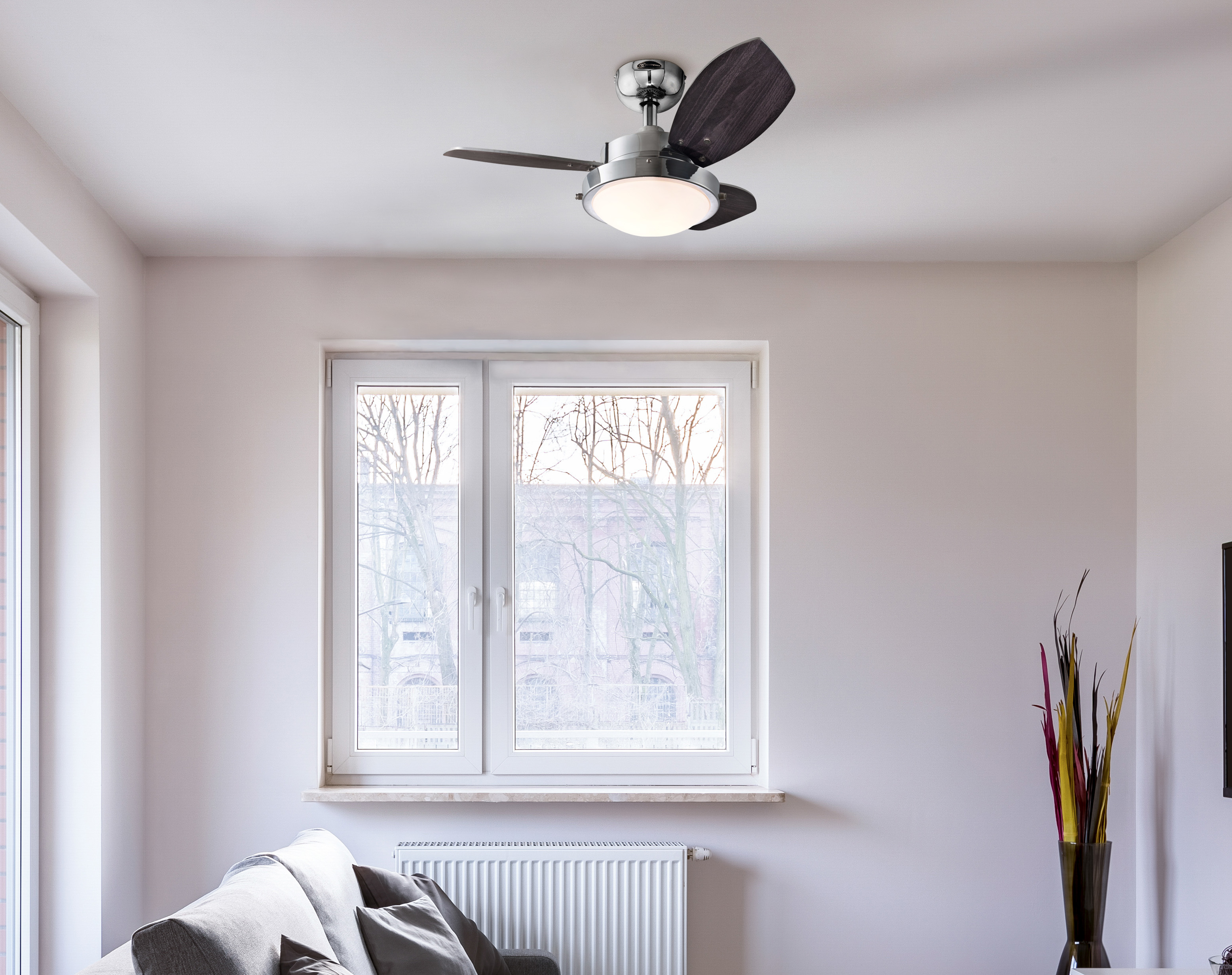 Ein Deckenventilator, ein Fenster mit Bäumen, ein Sofa und eine bunte Dekopflanze in einer Vase.