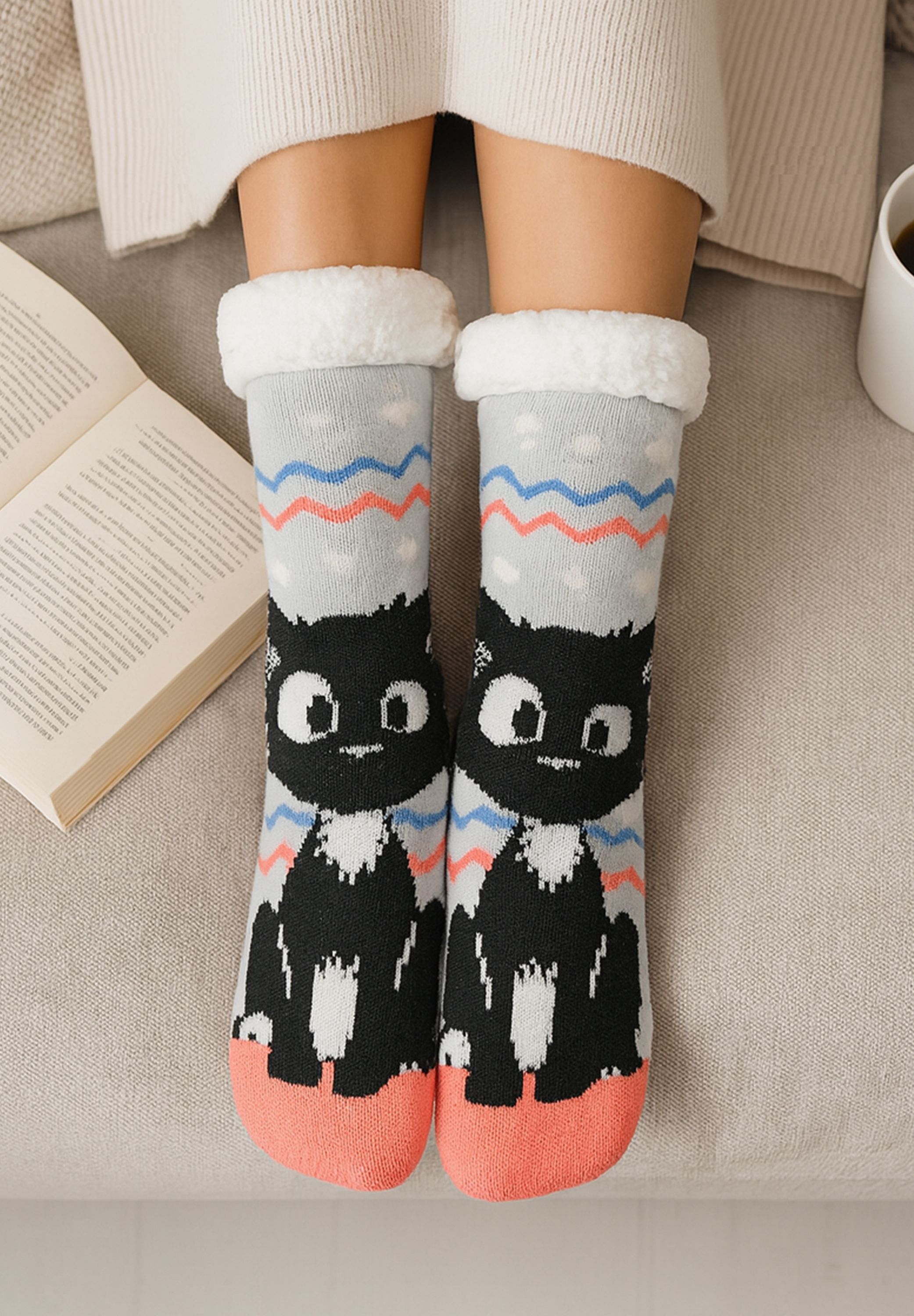 Person trägt Katzensocken. Offenes Buch und Tasse auf beigem Sofa. Socken haben schwarze Katzen, blaue und rote Zickzackmuster.