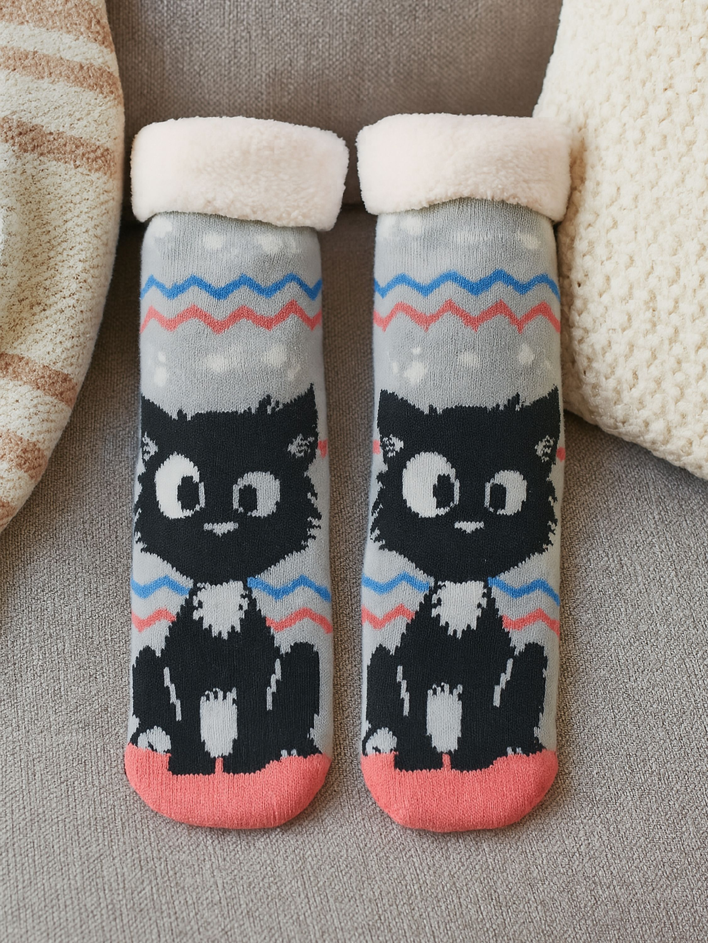 Paar Socken mit Katzenmotiv, weißem Besatz, grauem Körper, schwarzem Katzengesicht, rosa Zehen auf grauer Couch.