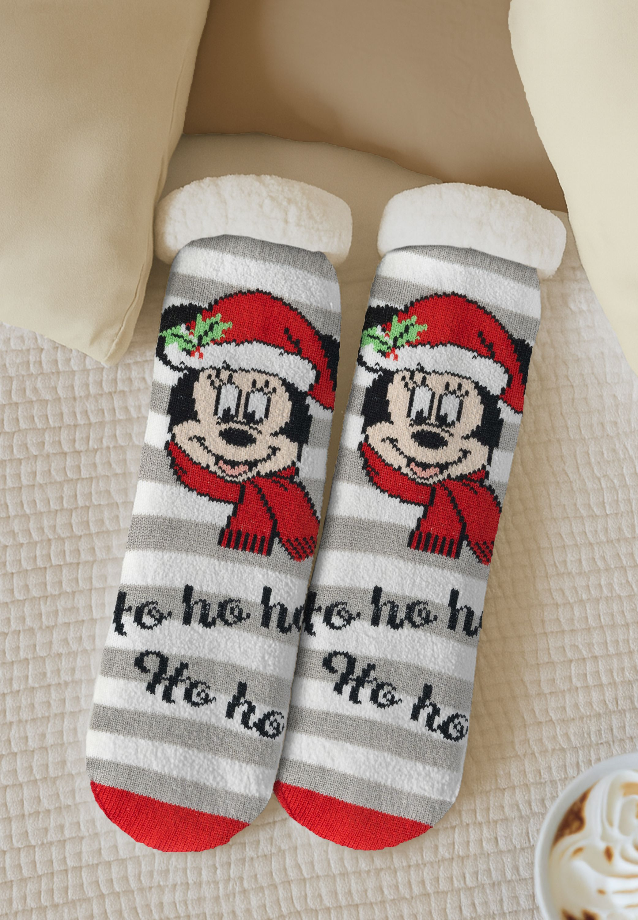 Zwei Weihnachtssocken mit Mickey Mouse. Sie haben Streifen, rote und weiße Details. Der Hintergrund zeigt Kissen und eine Tasse.