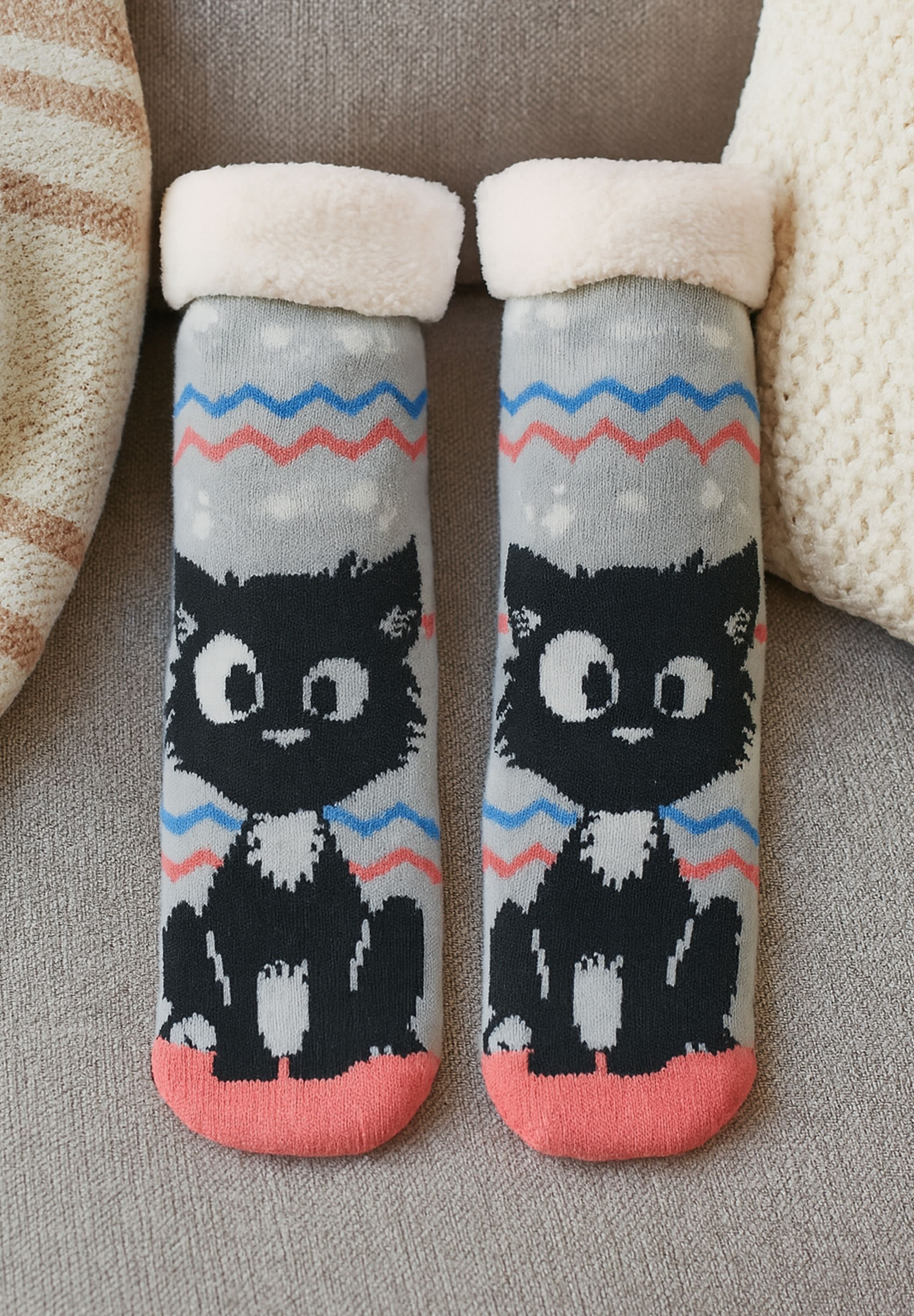 Paar Katzensocken mit grauen, blauen, roten Mustern, weißem Flaum oben und rosa Zehen, auf einem grauen Sofa.