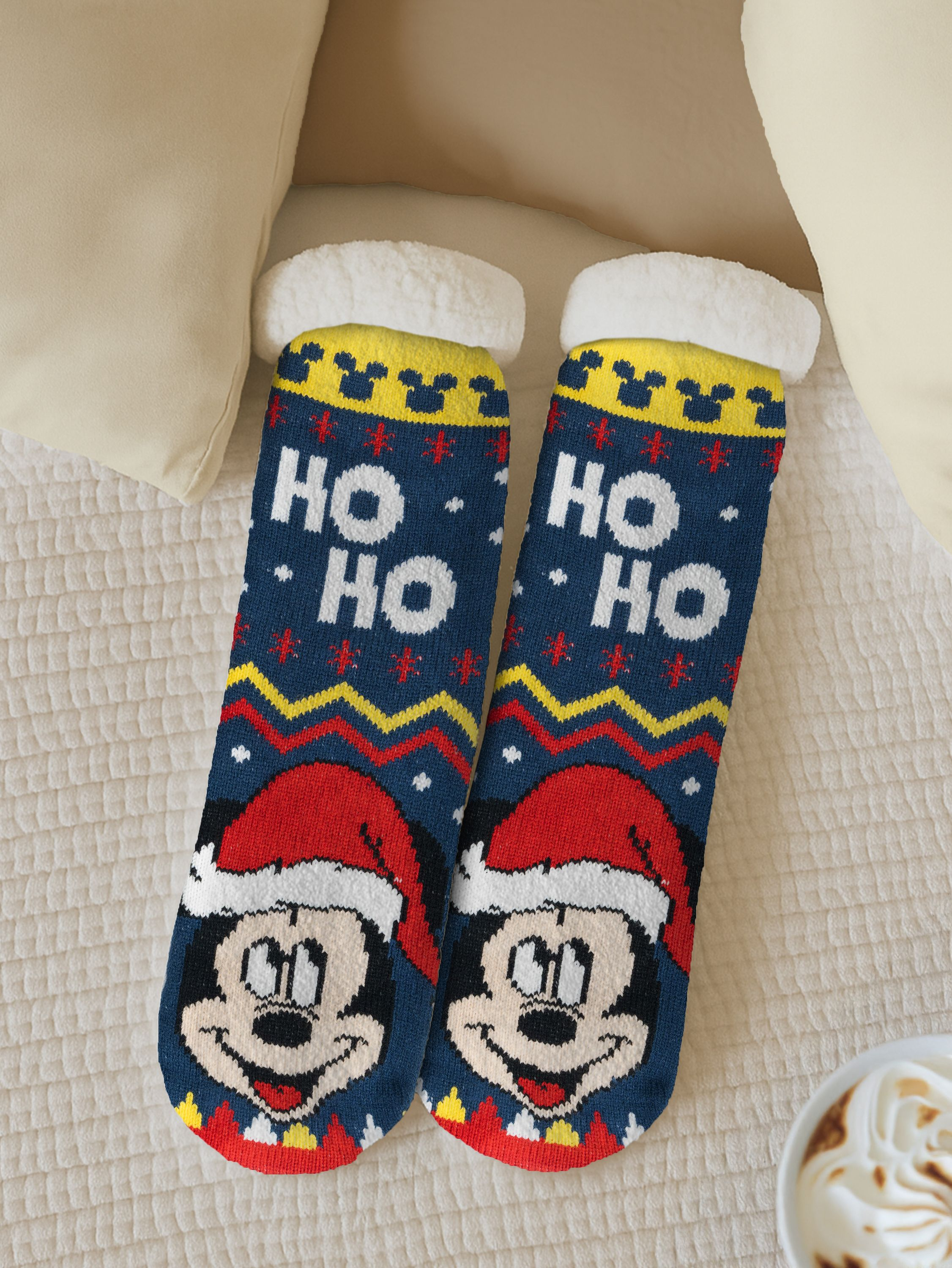 Zwei blaue Weihnachtssocken mit Mickey Mouse-Gesichtern, HO HO-Text und roten Weihnachtsmützen auf einem Bett mit Kissen und Kaffee.