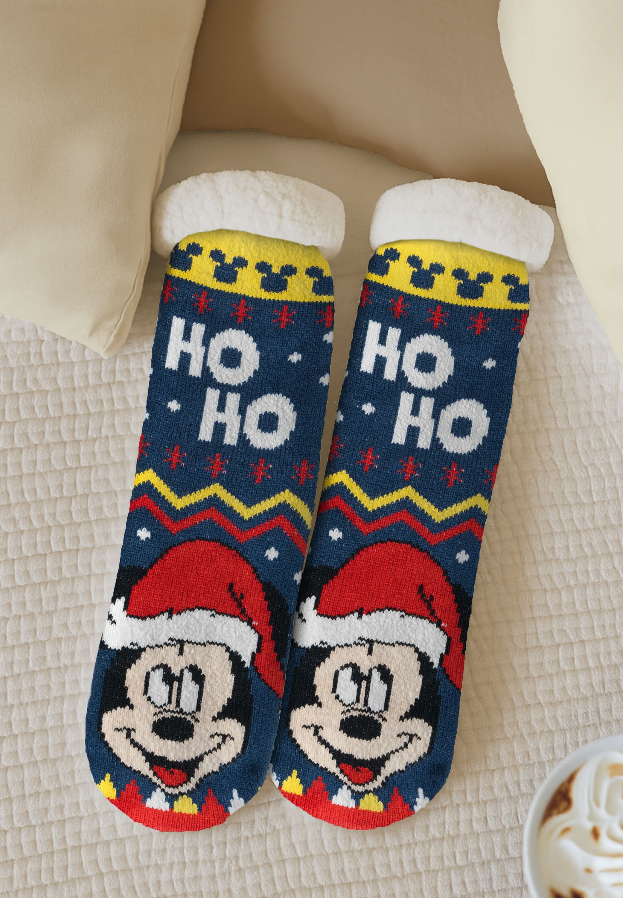 Zwei festliche Socken mit Mickey Mouse-Gesichtern und "HO HO"-Text auf einem Bett. Cremefarbene Kissen und eine Kaffeetasse sind sichtbar.