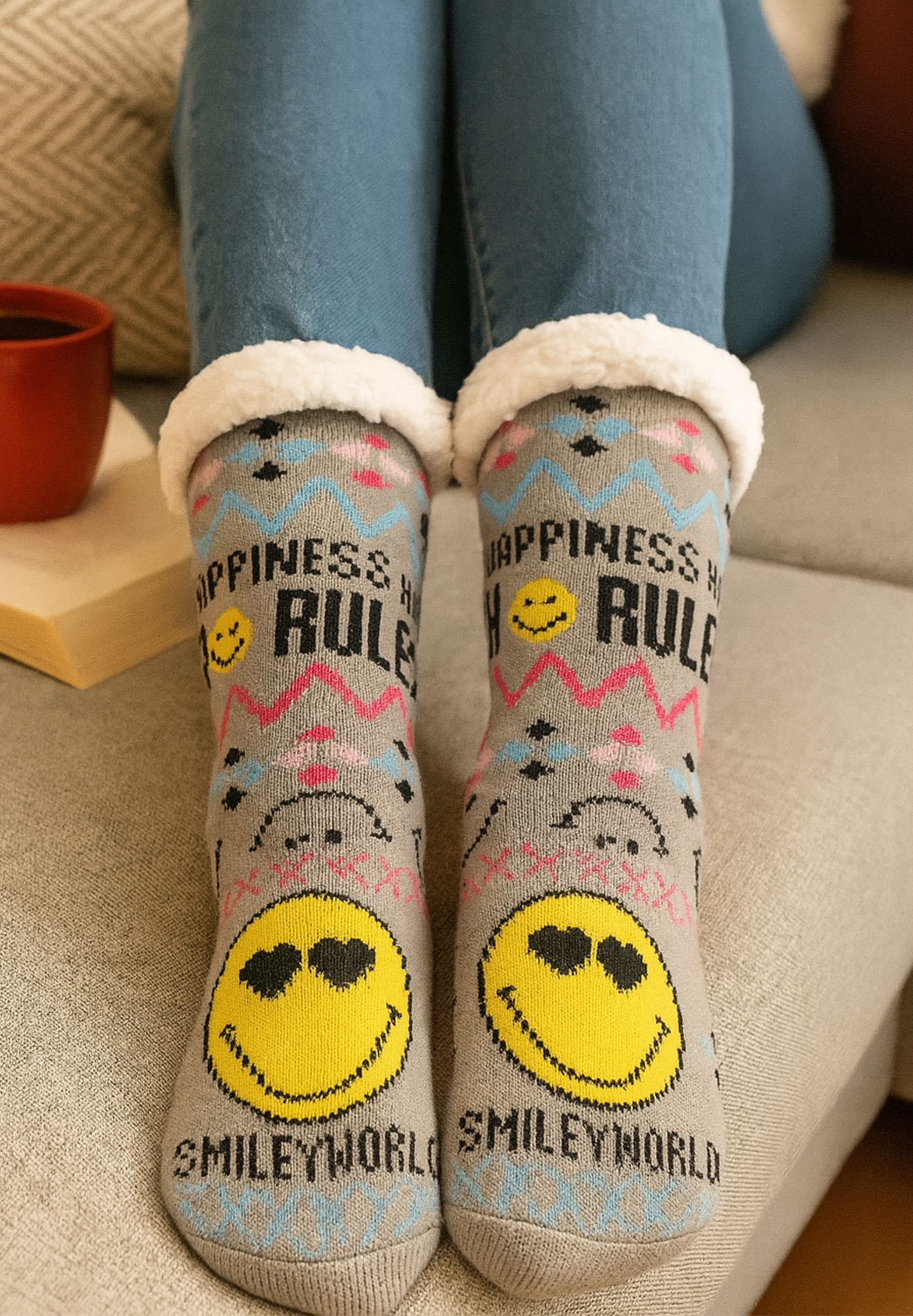 Person trägt graue Socken mit Smiley-Gesichtern und Text. Blaue Jeans sichtbar. Eine rote Tasse und ein Buch sind im Hintergrund.