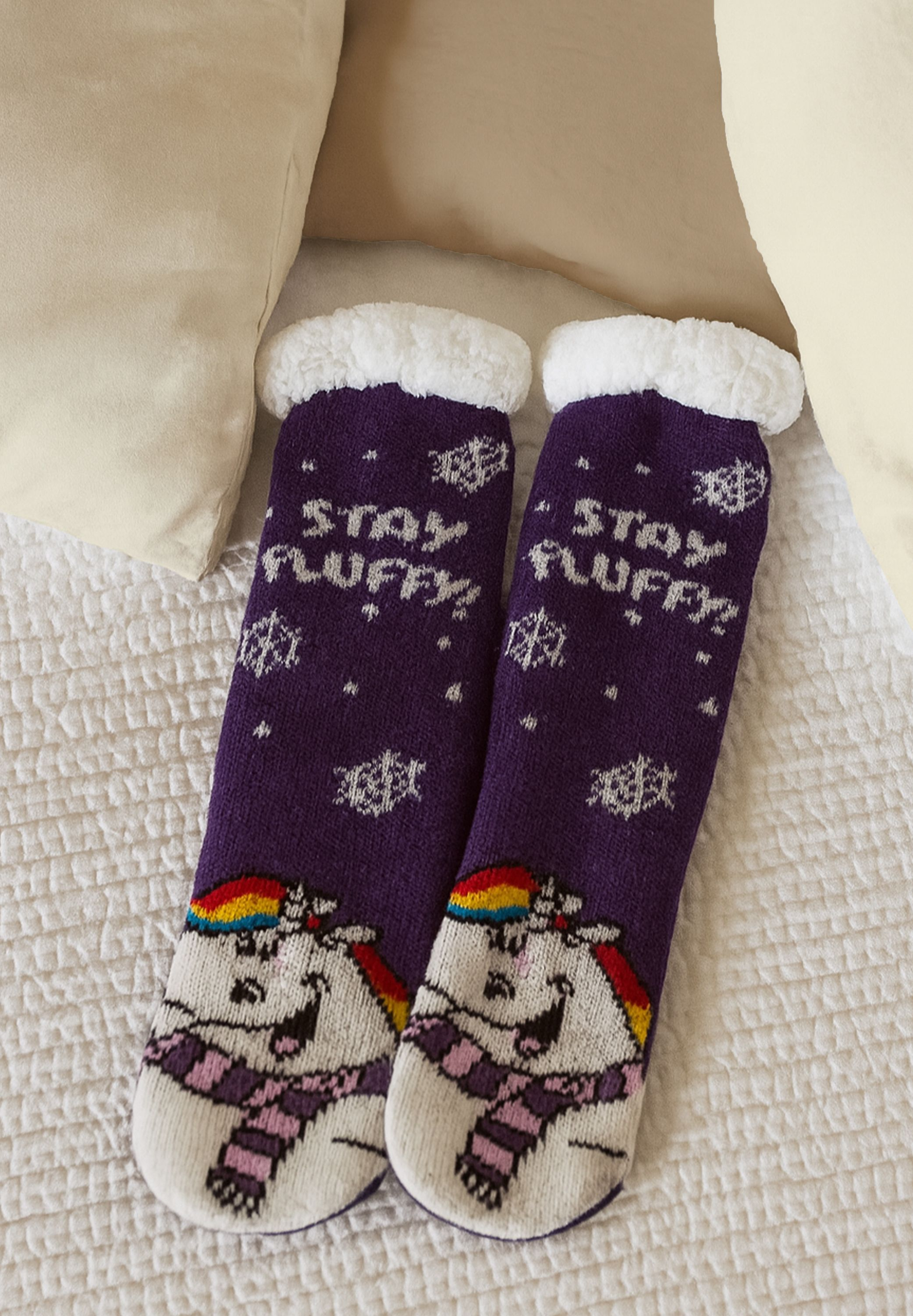Zwei violette Socken mit Einhorn-Design, dem Schriftzug 'Stay Fluffy' und Schneeflocken, auf einem weißen Bett.