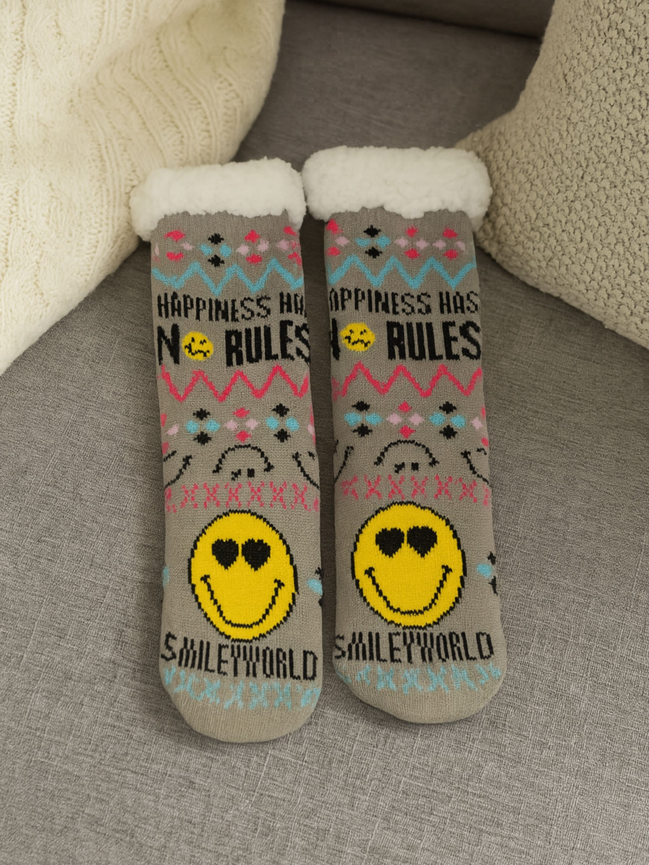 Zwei graue Socken mit Smiley-Gesichtern und dem Text 'Happiness has no rules' auf grauer Oberfläche, mit einer weißen Decke und Kissen.