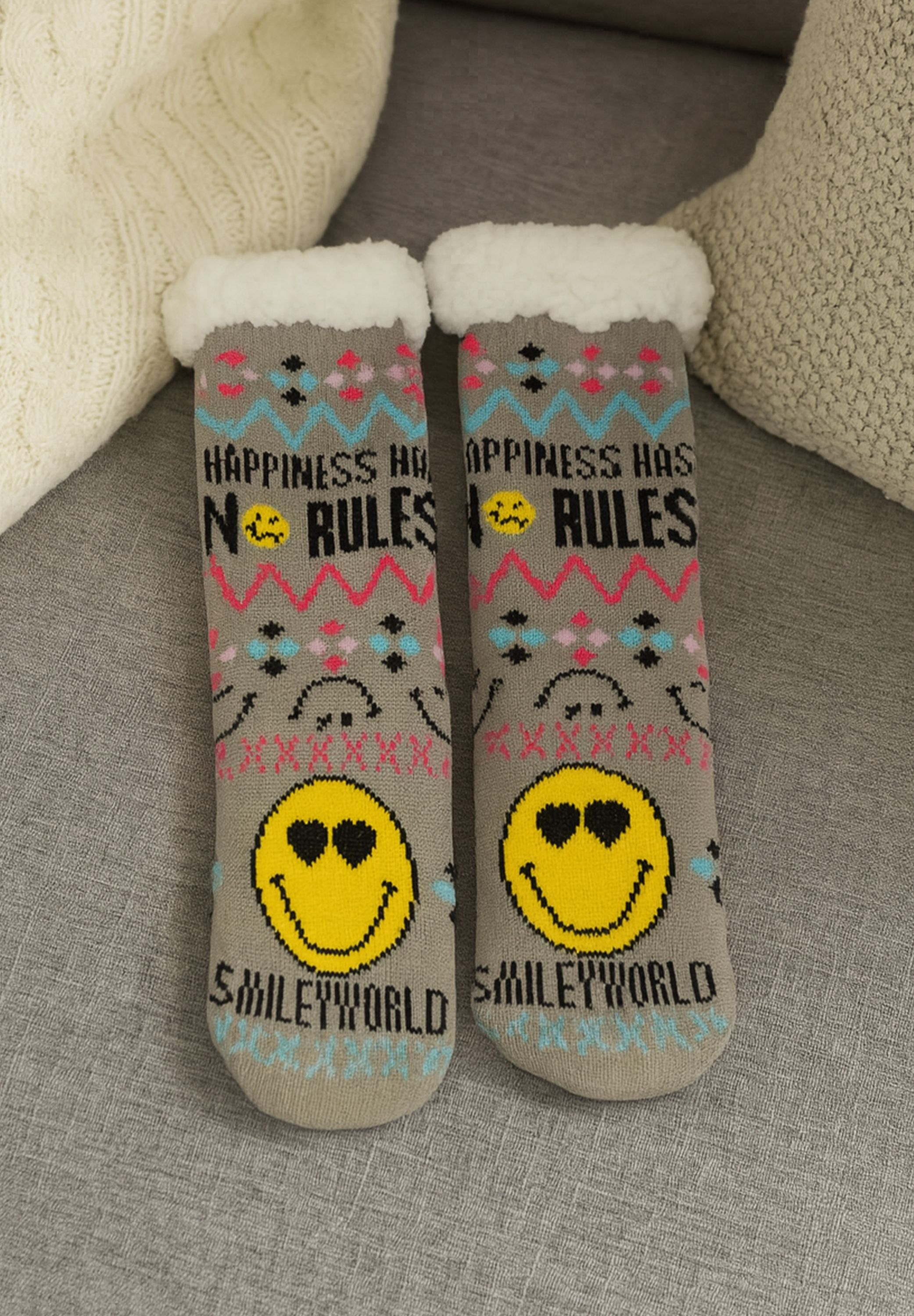 Zwei graue Socken mit Smiley-Gesichtern und dem Text "Happiness has no rules" auf einem grauen Sofa mit Kissen.
