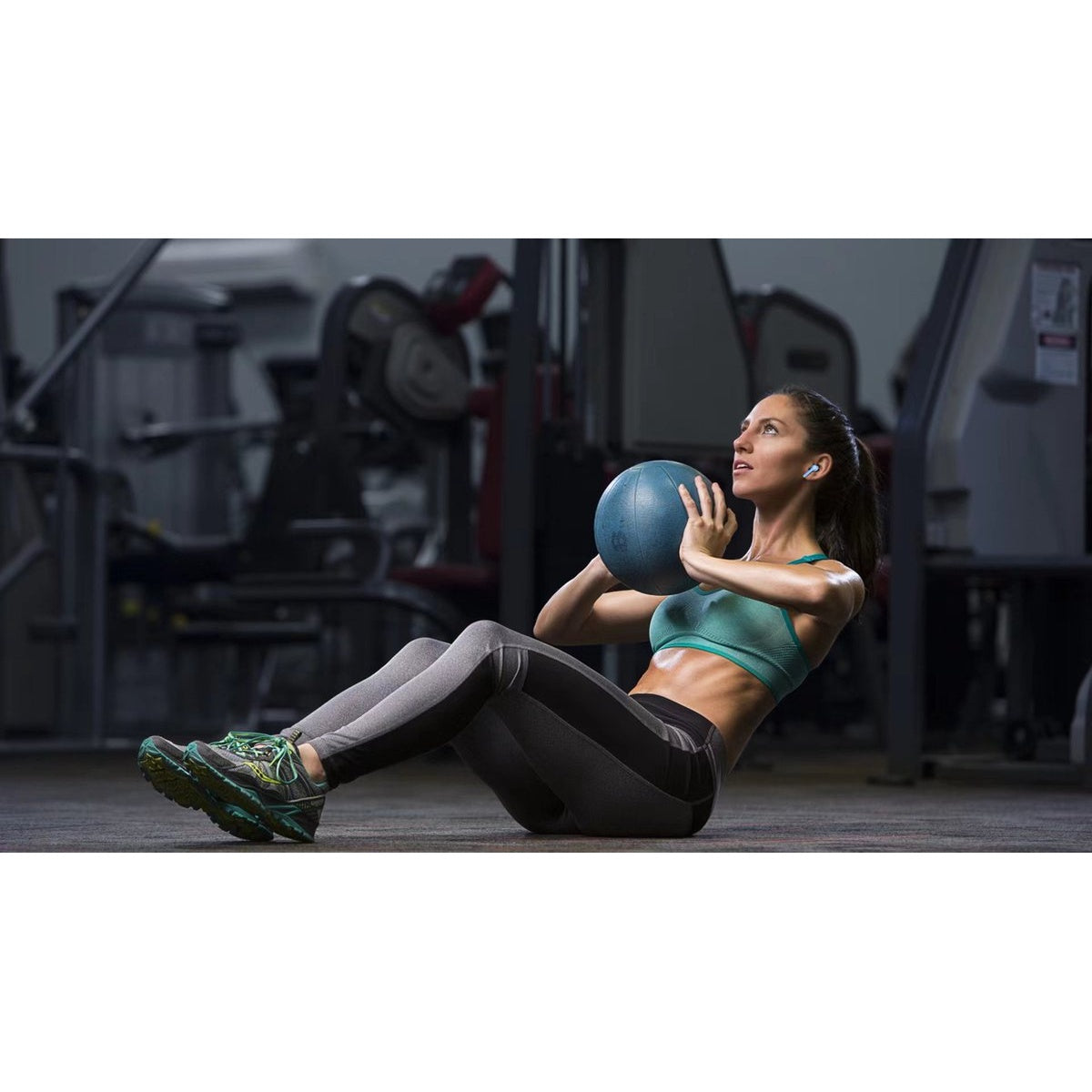 A woman in a gym does sit-ups, holding a blue ball. She wears sports attire. Gym equipment is visible in the background.