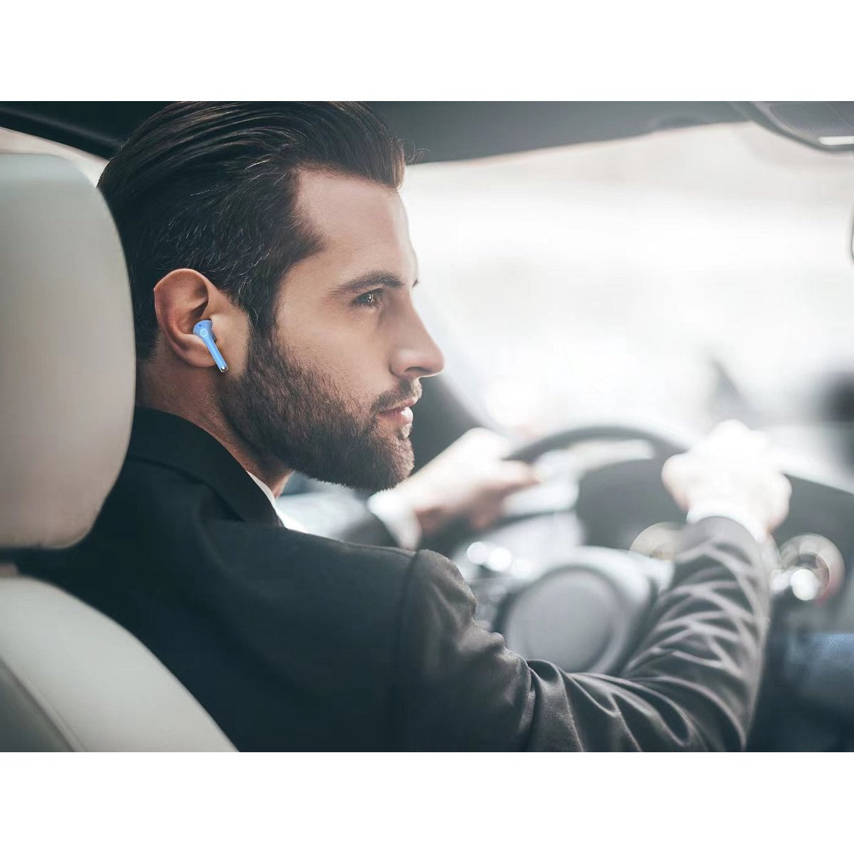 A man in a suit drives a car, wearing a blue earbud. He's looking ahead, holding the steering wheel. The focus is on his face.