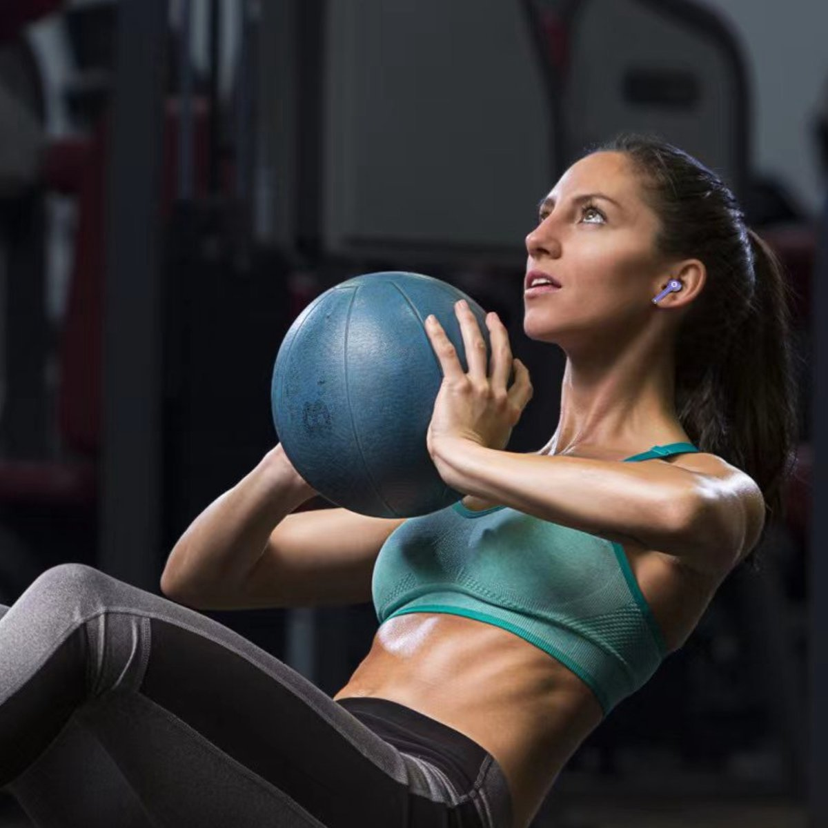 A woman in workout clothes is holding a blue medicine ball, looking up. She is in a gym. 