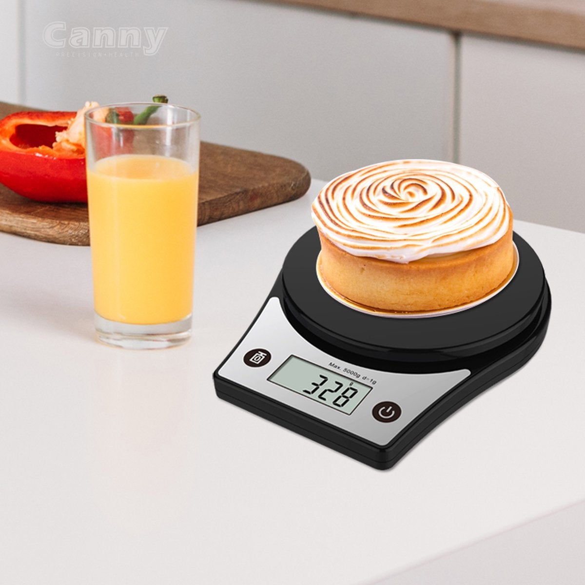 A black kitchen scale displays 328g with a cake on top. A glass of juice and a pepper are in the background.