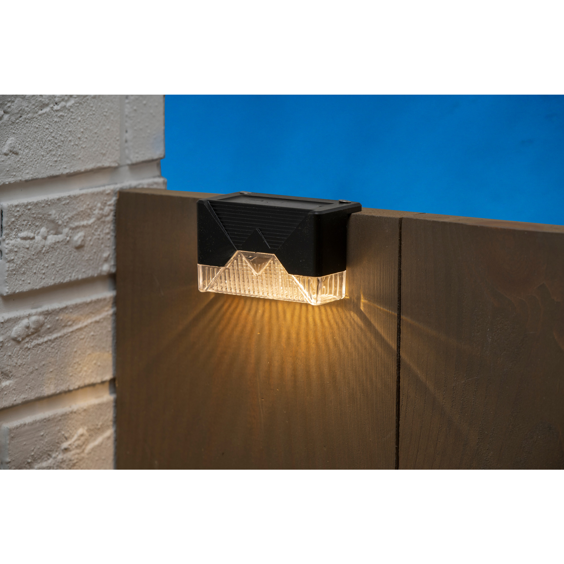 A solar-powered black and clear light fixture is mounted on a wooden fence, casting a warm glow against a blue sky.