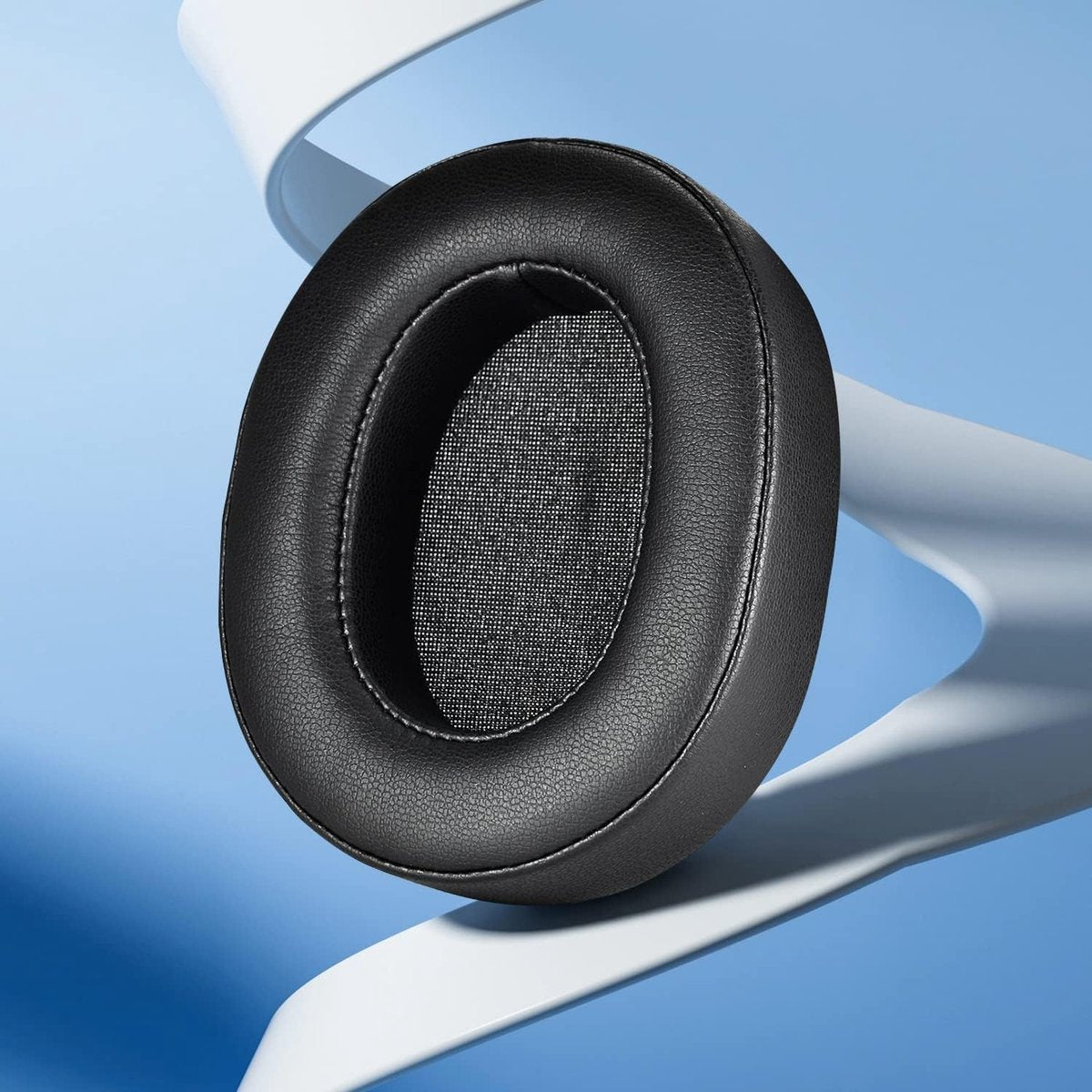 A black, oval-shaped headphone ear cushion floats. The interior is mesh, and the exterior is leather. A white curved shape is in the background.