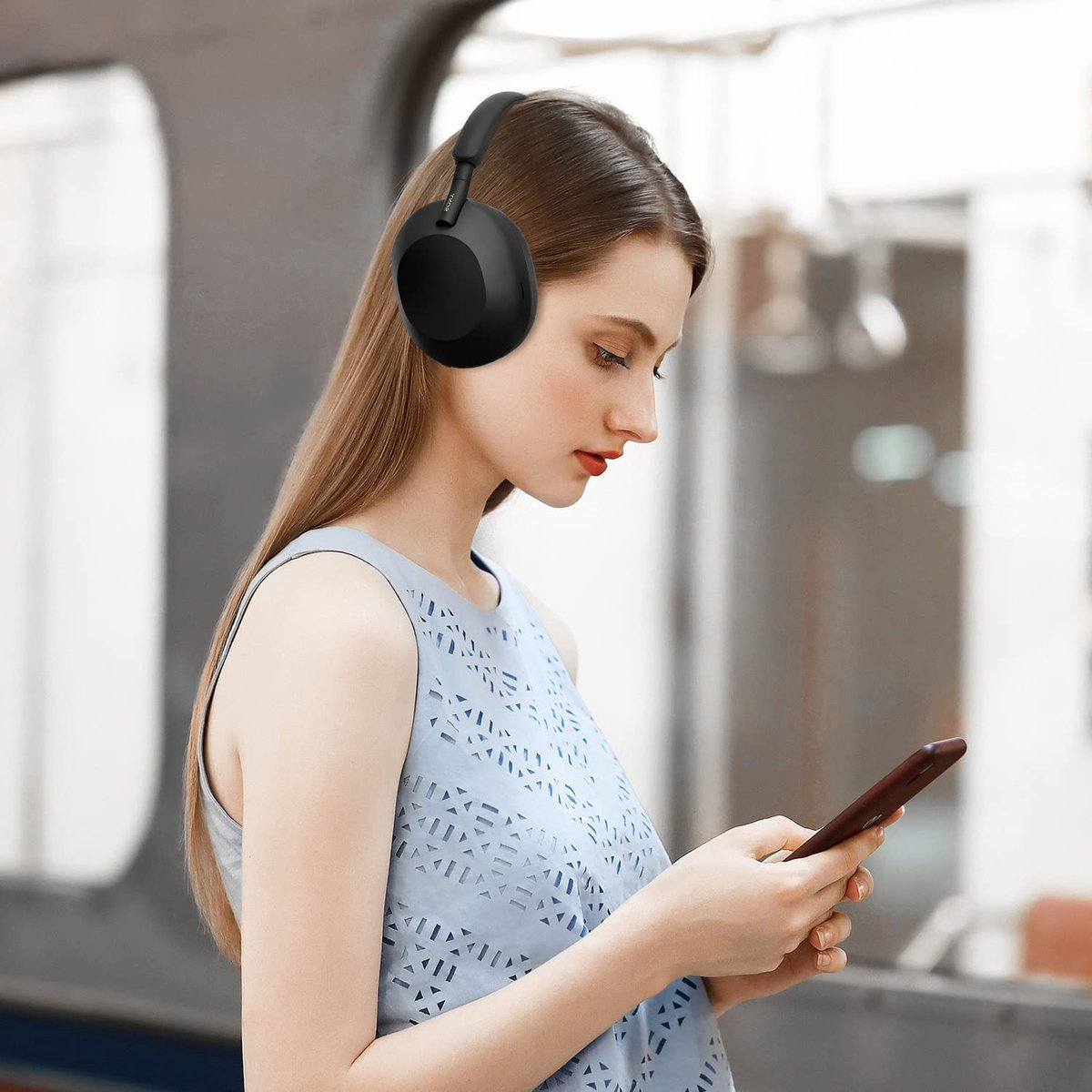 Woman wearing headphones, looking at a phone. She is wearing a blue top. The background is blurred.