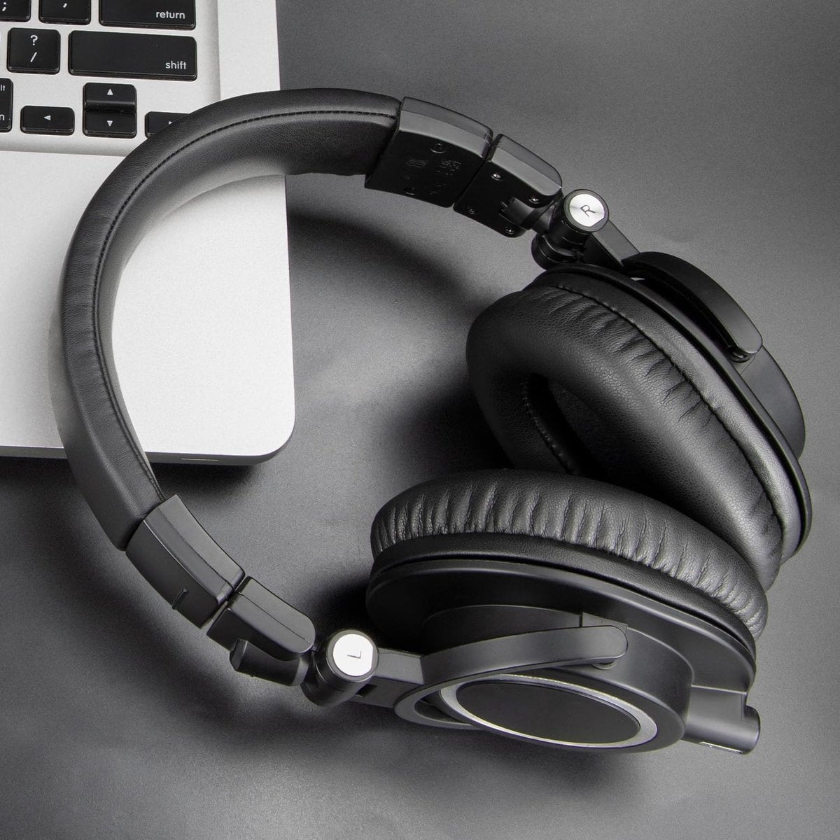 Zwarte koptelefoon ligt op een zwart oppervlak in de buurt van een laptop. De koptelefoon is over-ear. De achtergrond is donkergrijs.