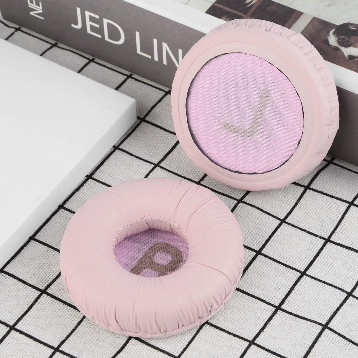 Twee roze oorkussens voor koptelefoons op geruite stof. Eén kussen is compleet, de andere heeft een centraal gat. Boeken op de achtergrond.