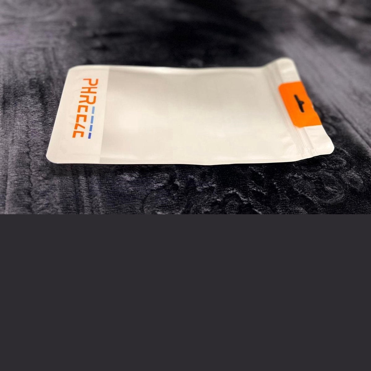 A white bag with 'PHREECLE' printed on it, orange seal, on a dark, textured surface. Background is dark gray.