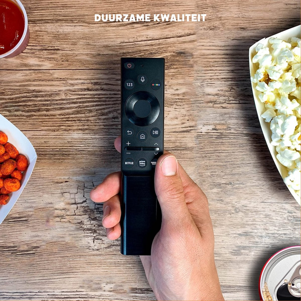 Hand holding a black remote control. Snacks, popcorn, and a drink are beside it on a wooden table.