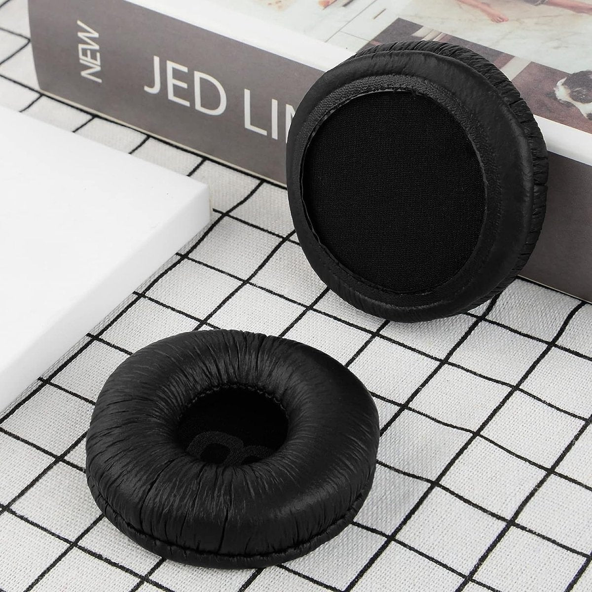 Twee zwarte oorkussens voor hoofdtelefoons op een geruit oppervlak, in de buurt van een boek met de titel 'JED LI' en een witte doos.