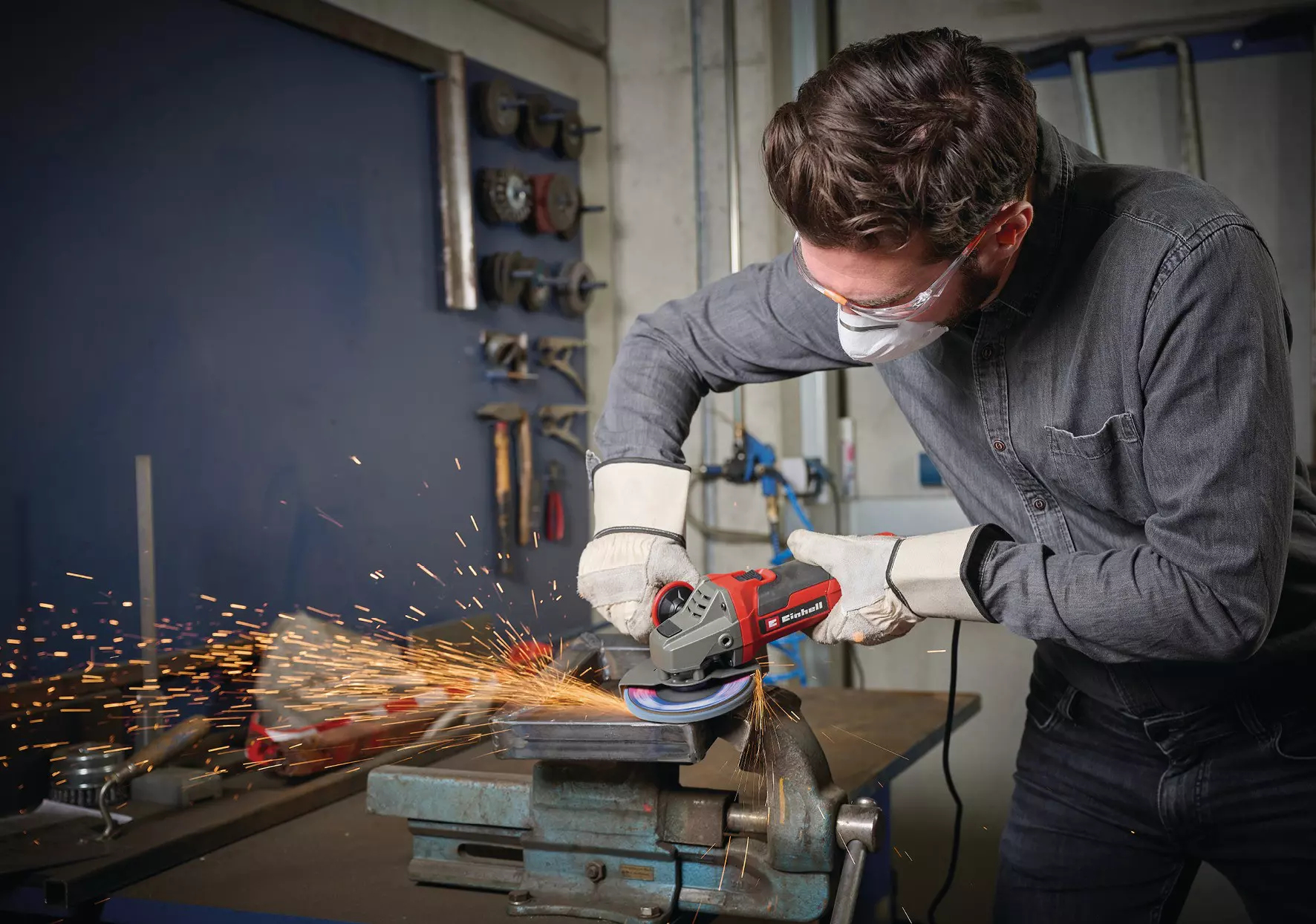 Mężczyzna szlifuje metal szlifierką kątową, lecą iskry. Ma okulary ochronne, maskę i rękawice w warsztacie.