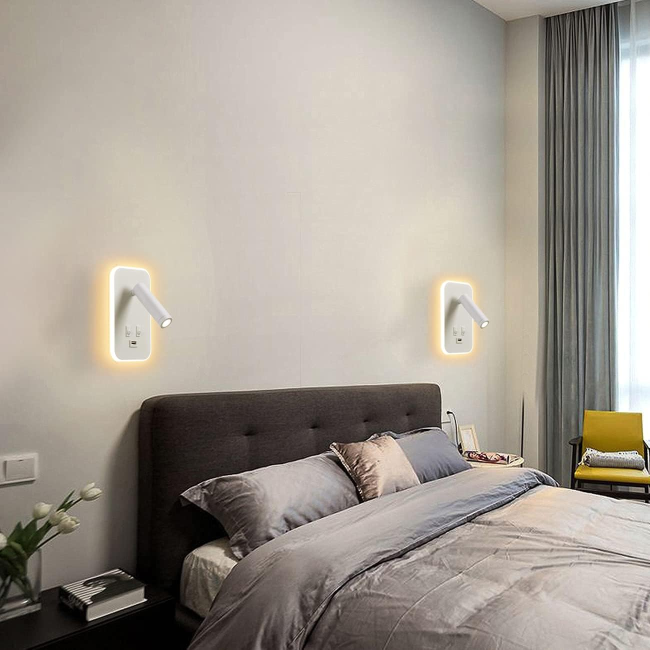 Schlafzimmer mit Bett, Lampen und Vorhängen. Zwei Wandlampen mit Lichtern. Graue Bettwäsche und Kissen. Gelber Stuhl.