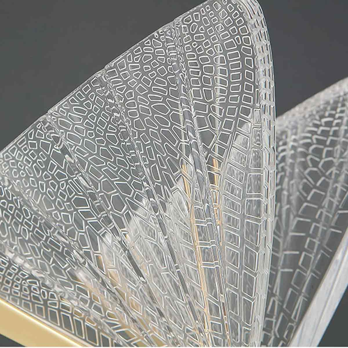 Nahaufnahme eines durchsichtigen, gemusterten Objekts mit Goldrahmen, möglicherweise einer Lampe. Das Objekt hat geometrische Muster und ist vor grauem Hintergrund.