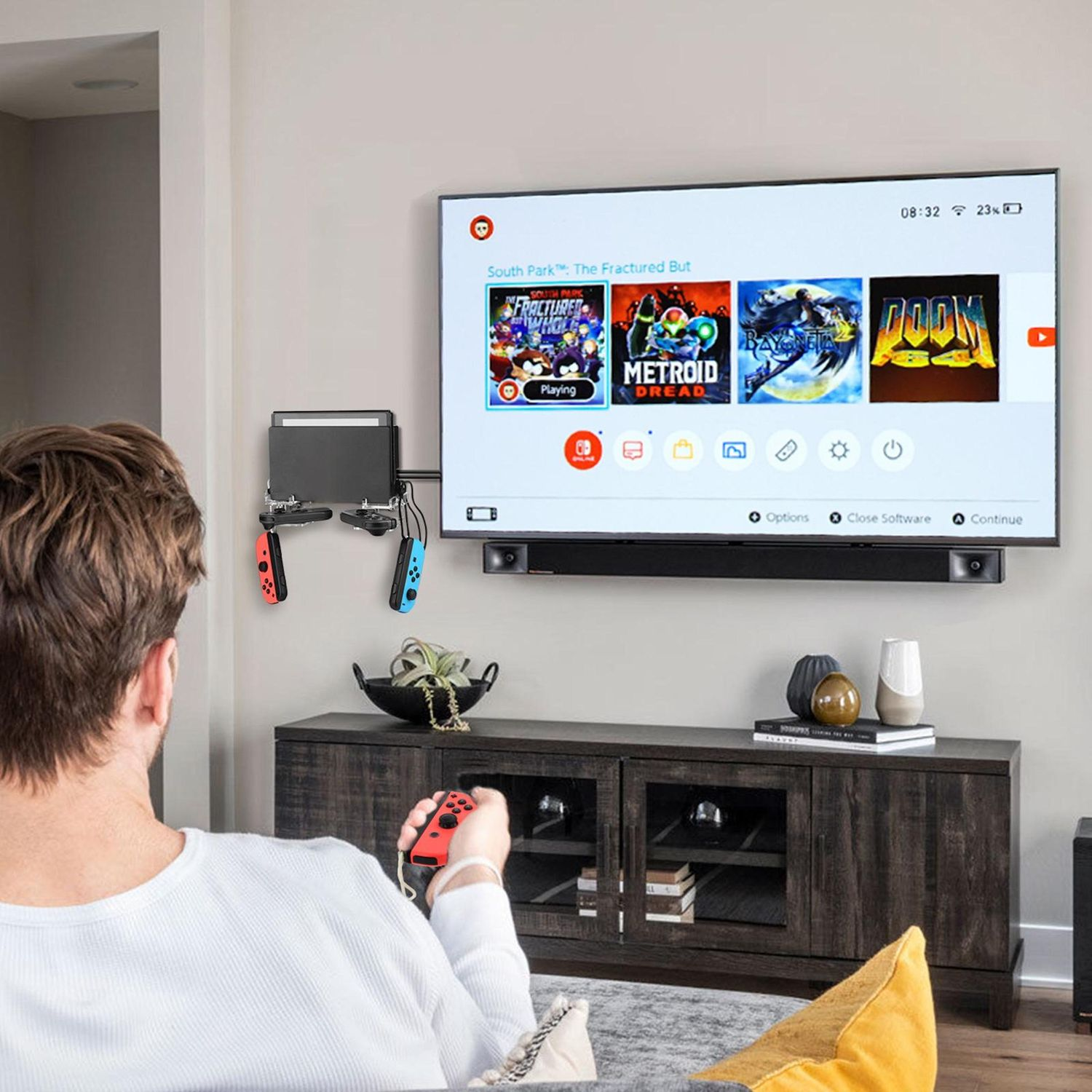 Eine Person hält einen roten Controller und spielt ein Spiel auf einem Fernseher. Der Fernseher zeigt Spielsymbole an. Eine Spielkonsole ist an der Wand montiert.