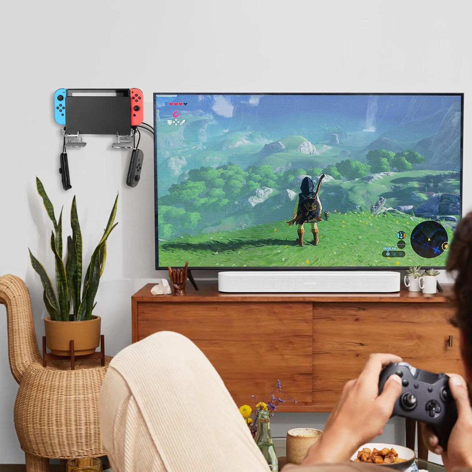 Person spielt Videospiel auf dem Fernseher. Eine Nintendo Switch ist an der Wand befestigt. Ein Korbstuhl und eine Pflanze befinden sich links.
