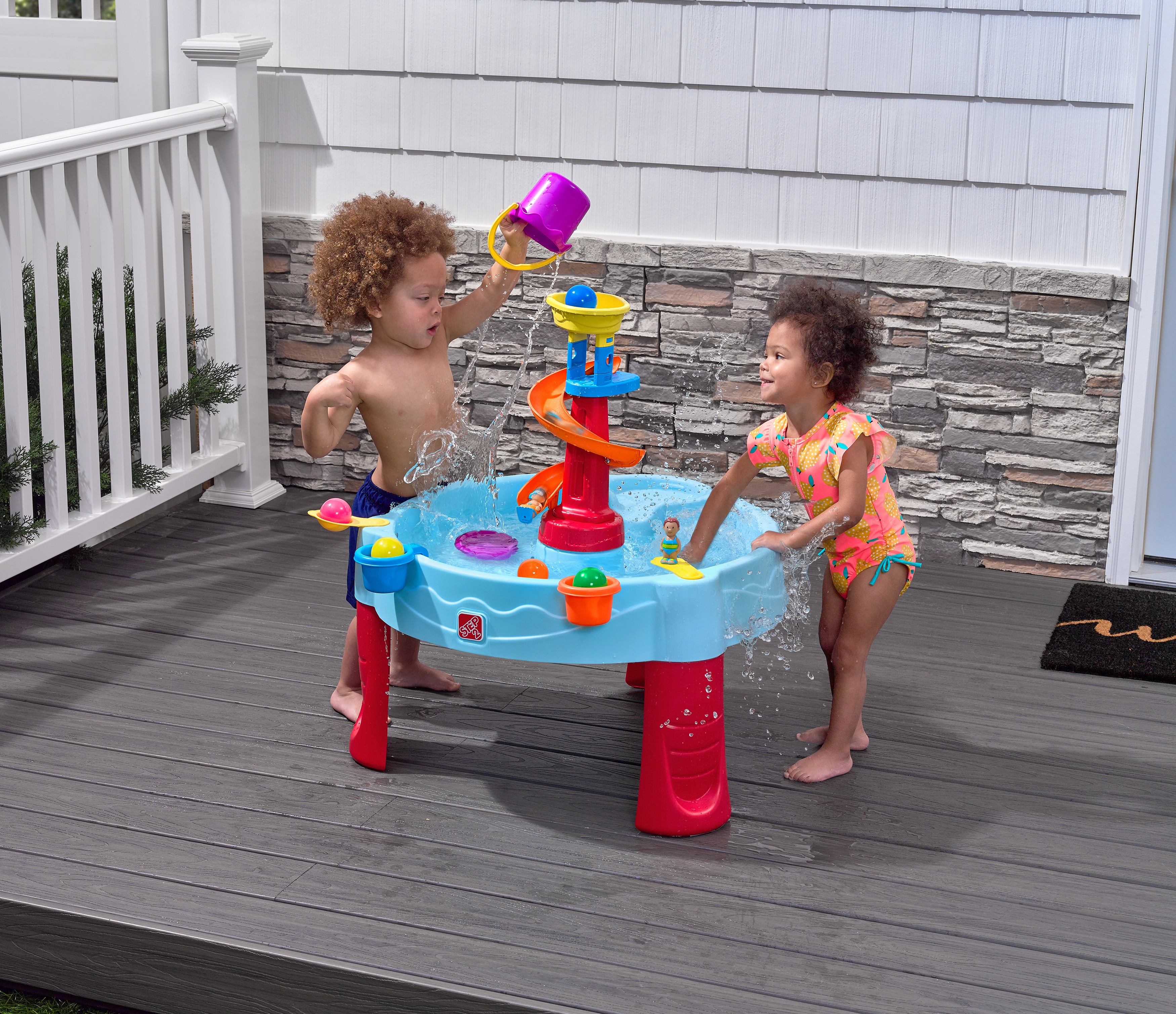 Zwei Kinder spielen mit einem Wassertisch. Einer gießt Wasser aus einem lila Eimer. Der andere berührt das Wasser.