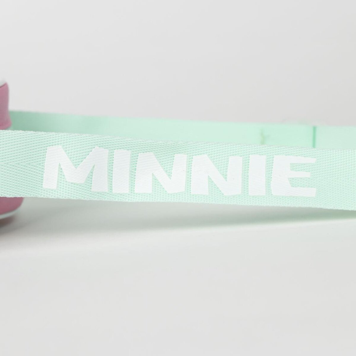 Una correa verde claro con la palabra 'MINNIE' en letras blancas grandes. El fondo es blanco liso.