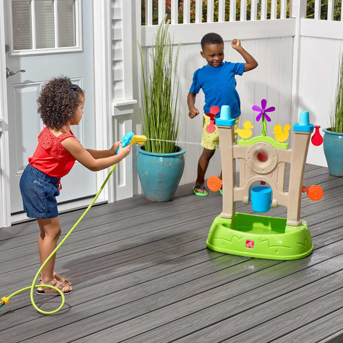 Zwei Kinder spielen mit Wasserspielzeug auf einer Terrasse. Ein Kind sprüht Wasser, das andere tanzt in der Nähe eines Wassertisches.