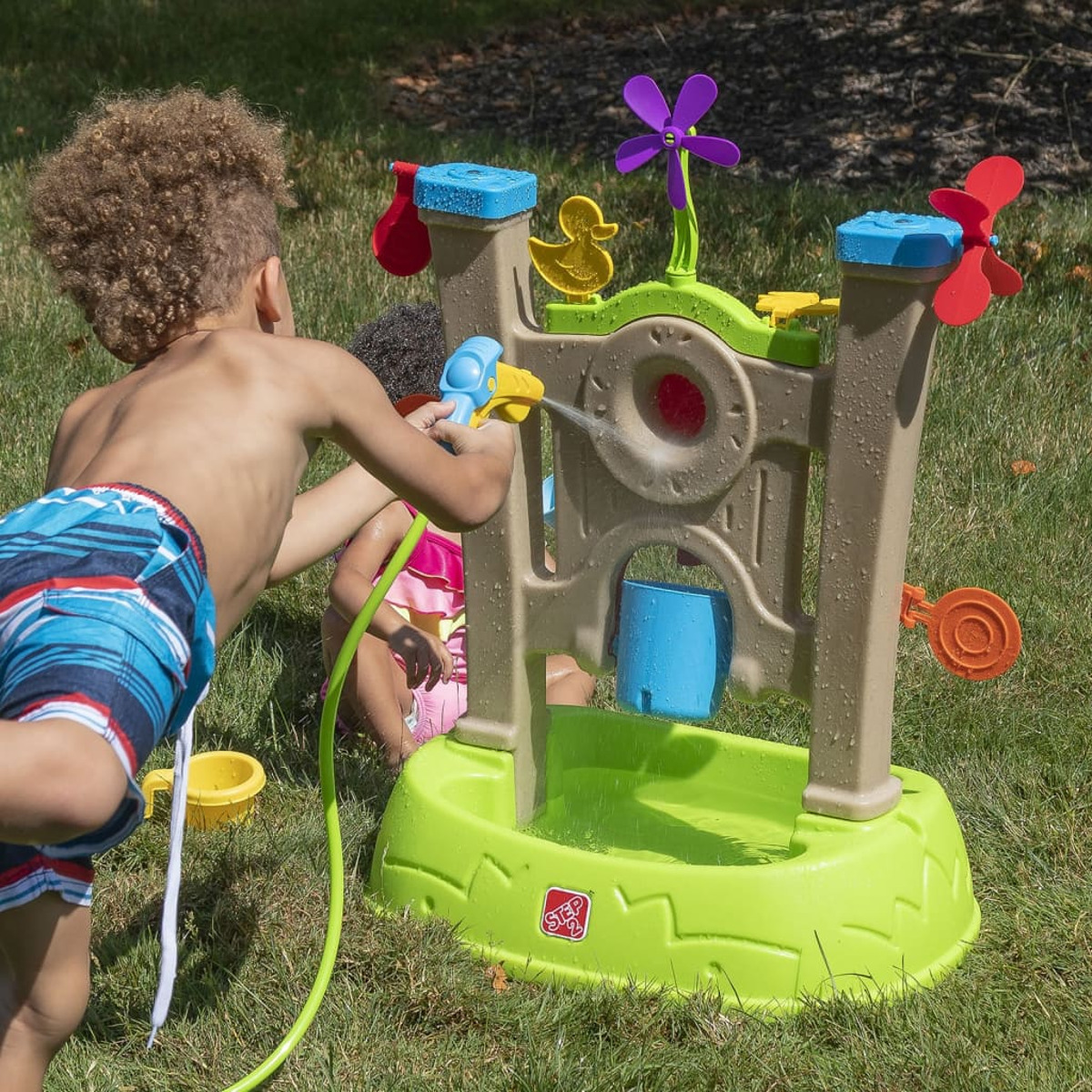 Zwei Kinder spielen mit einem Wasserspielzeug auf Gras. Eines sprüht Wasser. Das Spielzeug hat eine Ente, eine Windmühle und ein Wasserbecken.
