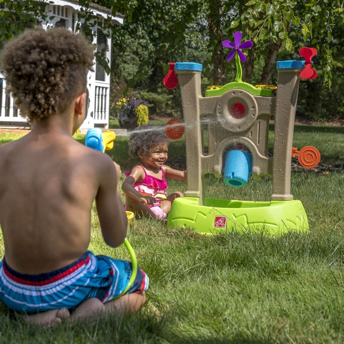 Zwei Kinder spielen mit einem Wassertisch auf dem Rasen. Ein Kind sprüht Wasser, das andere sitzt am Tisch.