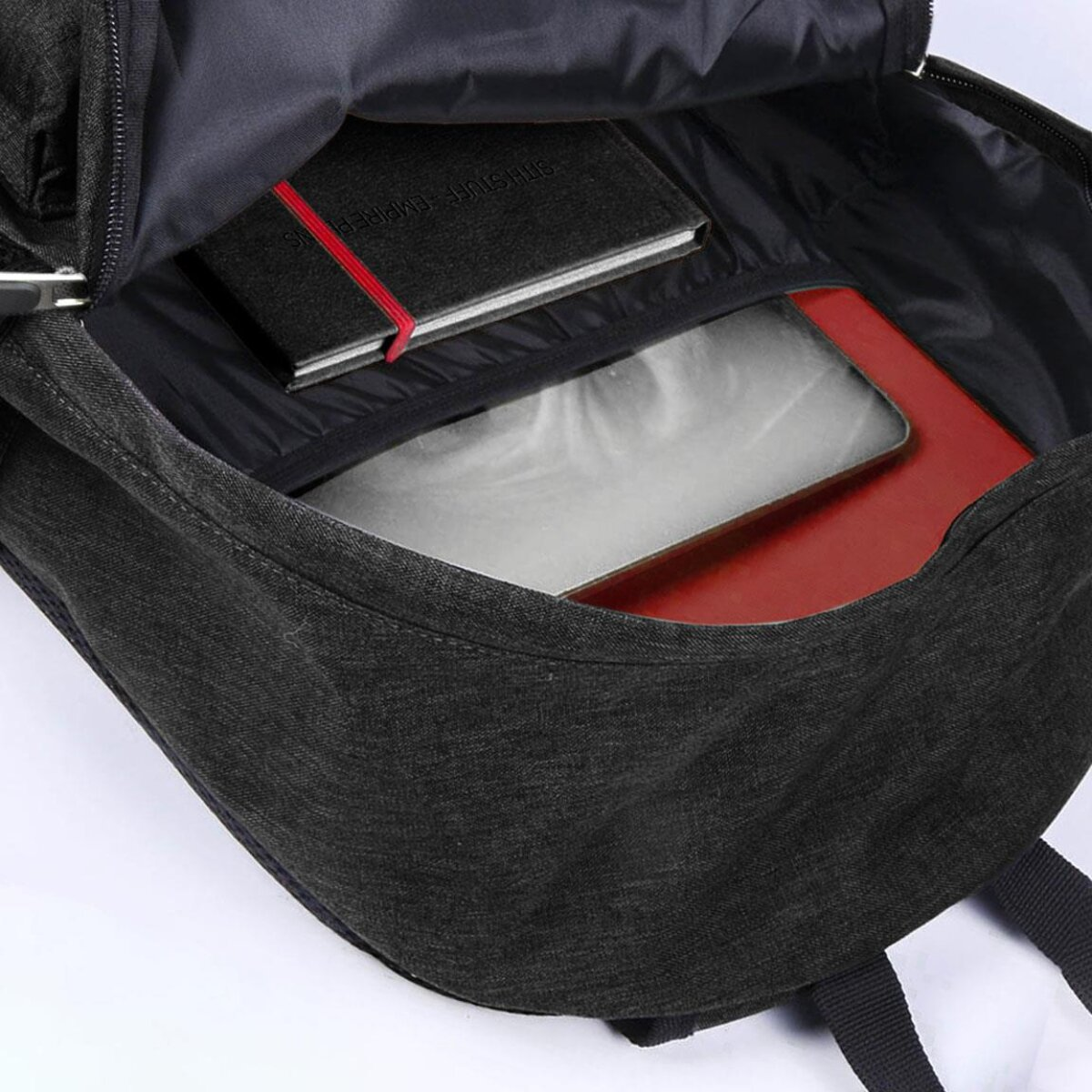 Dentro de una mochila negra: un cuaderno negro con una banda roja, una tableta plateada y un cuaderno rojo. La mochila está abierta.