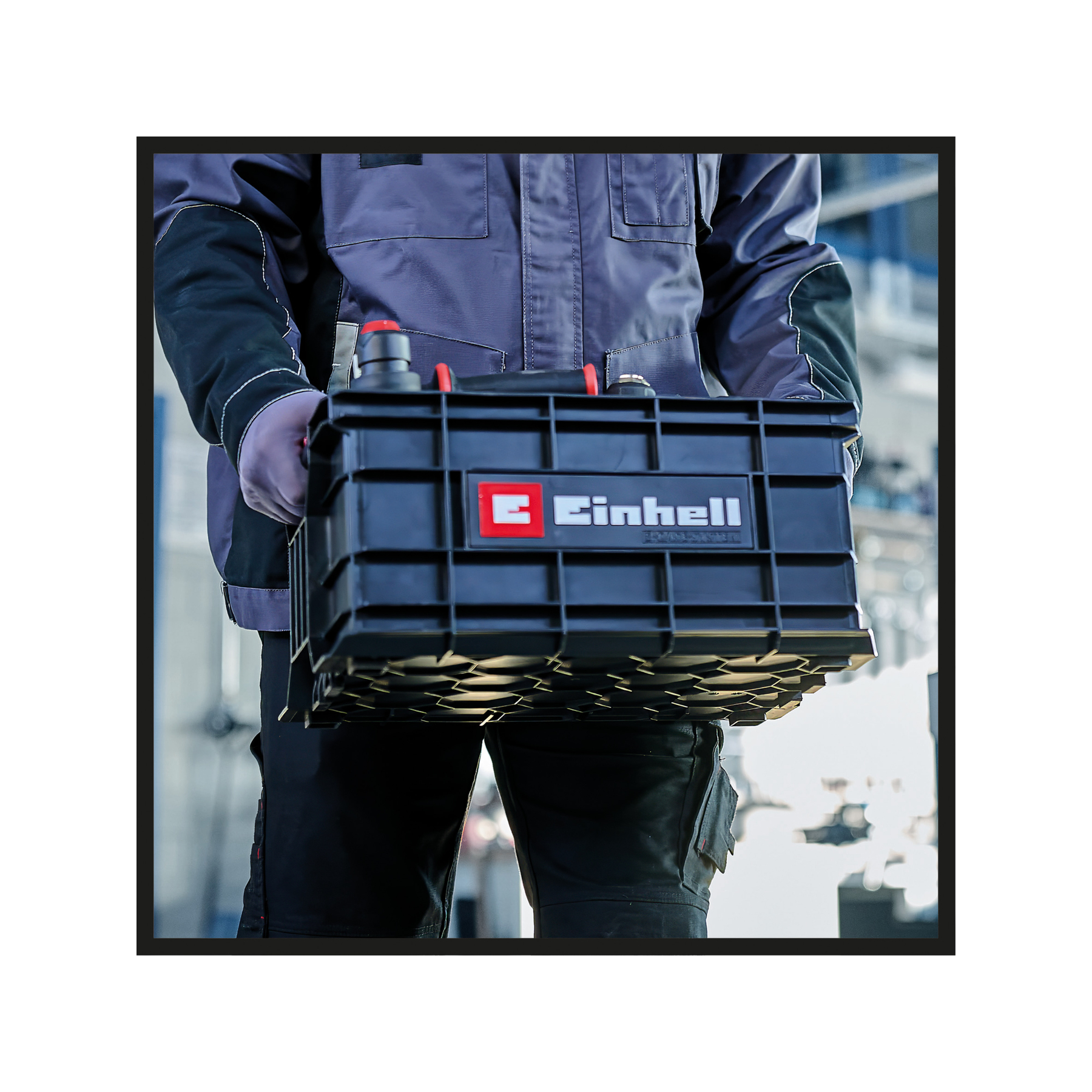 Eine Person hält eine schwarze Einhell-Werkzeugkiste. Die Person trägt eine graue Jacke und eine schwarze Hose. Der Hintergrund ist verschwommen.