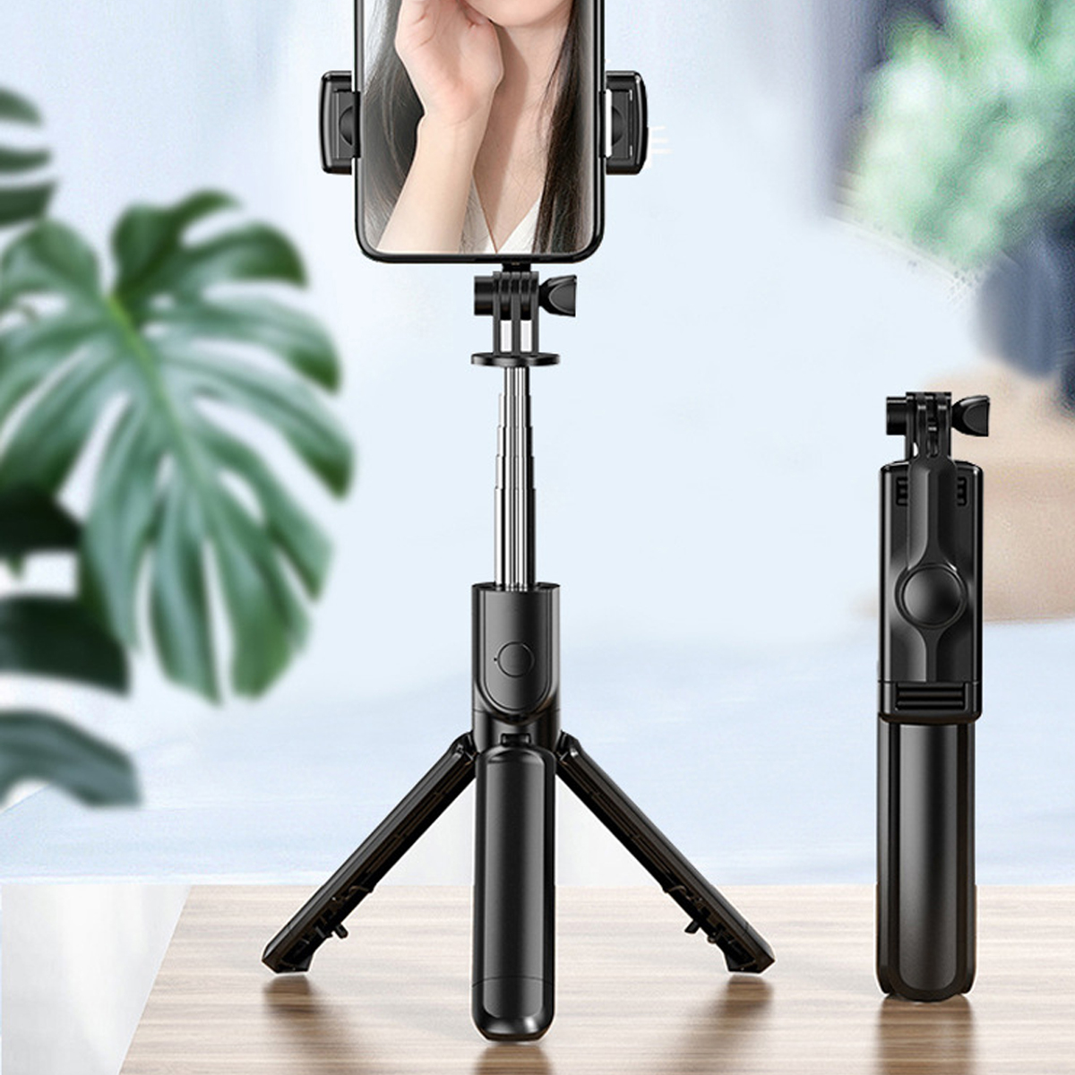 Ein schwarzer Selfie-Stick mit einem Telefon, das das Gesicht einer Frau zeigt. Ein abgetrennter Stick befindet sich rechts, eine grüne Pflanze unscharf im Hintergrund.