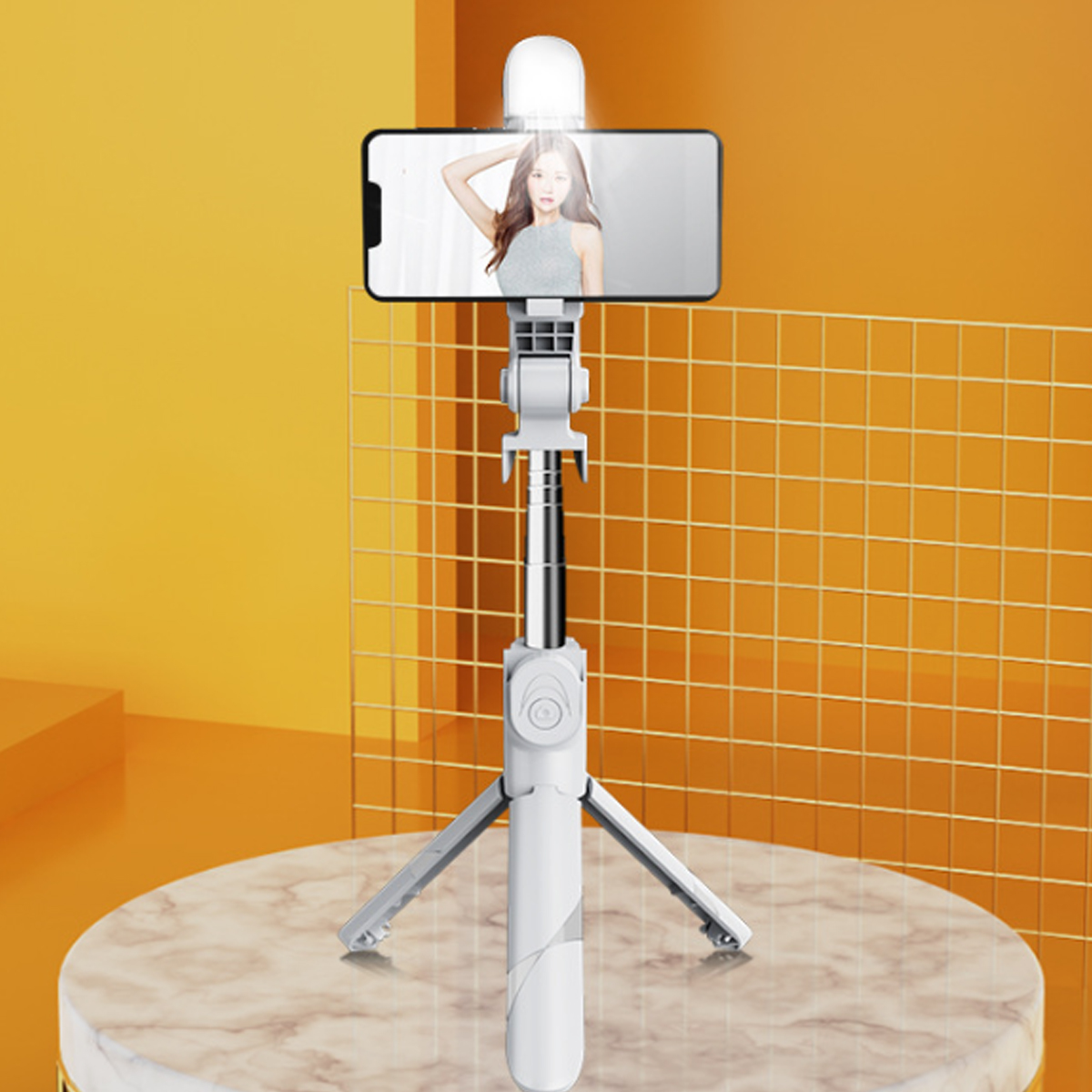 Ein Selfie-Stick mit einem Telefon, das das Bild einer Frau zeigt, vor einem orangefarbenen Hintergrund mit Gittermuster und Marmorständer.