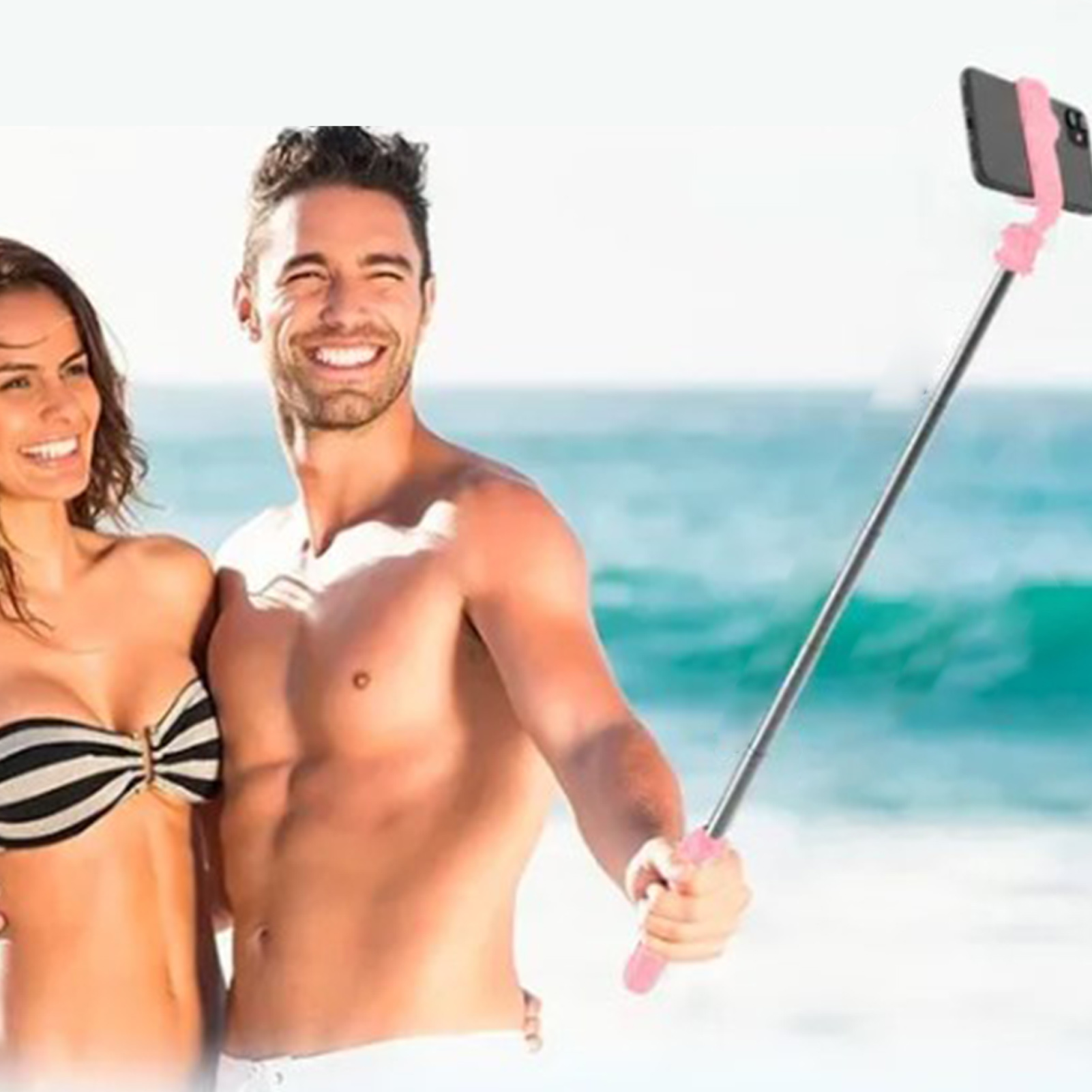 Ein Paar lächelt in die Kamera, ein Mann hält einen Selfie-Stick mit einem Telefon. Sie sind am Strand.