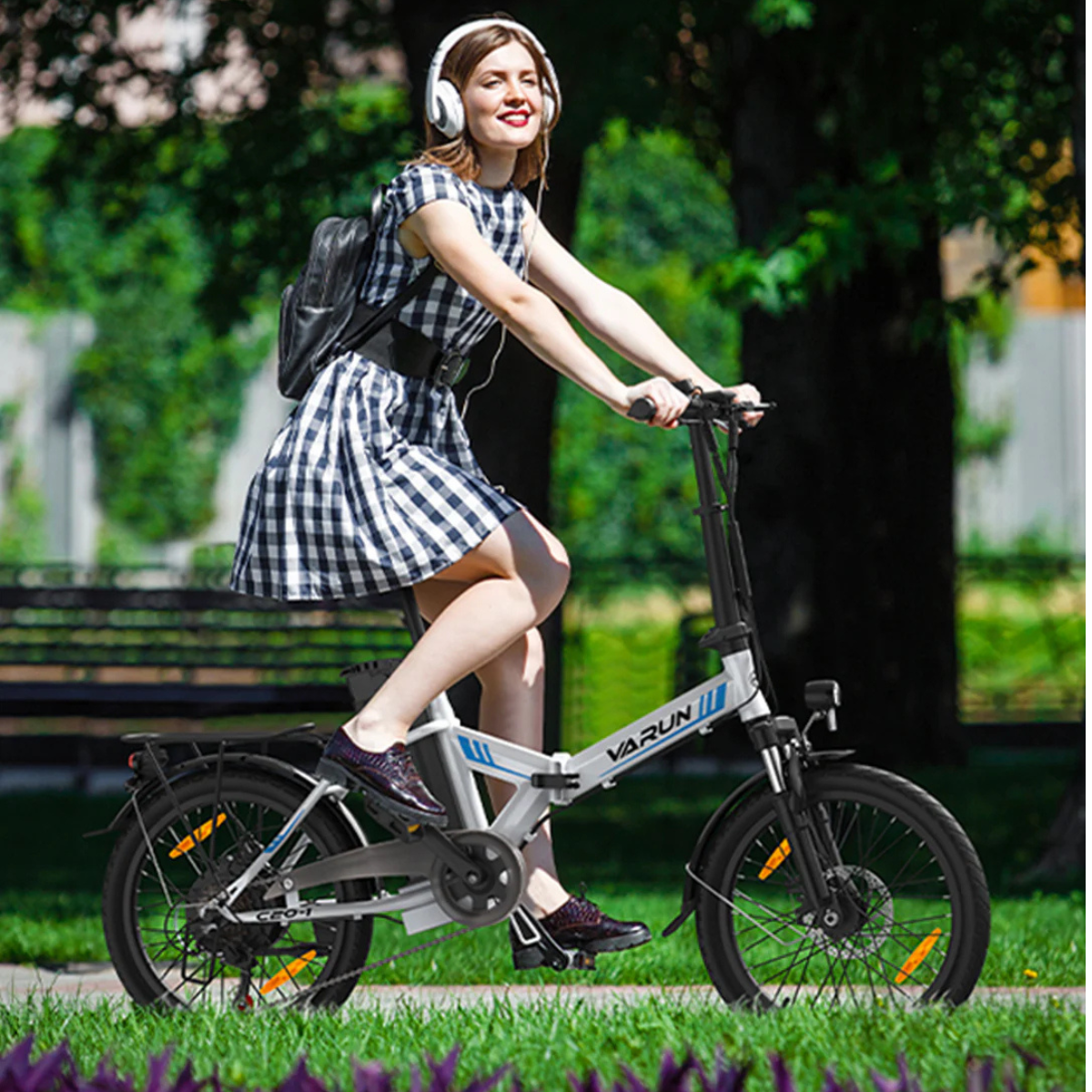 Kobieta na rowerze w słuchawkach, w szachownicowej sukience, z plecakiem, jadąca po trawiastym szlaku, z drzewami w tle.