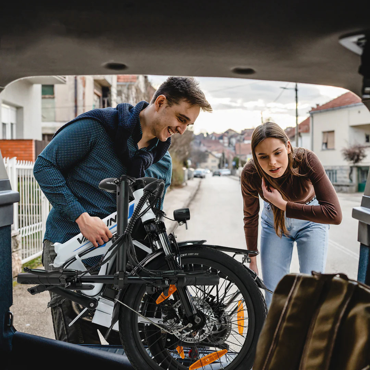 Mężczyzna i kobieta ładują złożony rower do bagażnika samochodu. Domy w tle wzdłuż ulicy. Kobieta ma na sobie dżinsy.