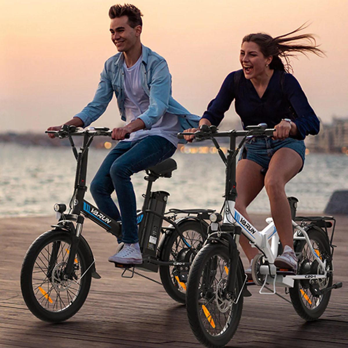 Dwie osoby jadą na trójkołowych rowerach po drewnianym pomoście. Uśmiechają się, ciesząc się jazdą. Tło to plaża i budynki.