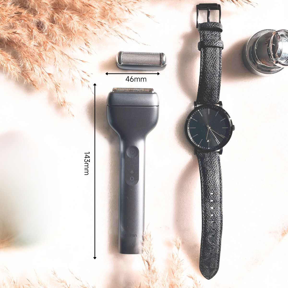 Een zwarte elektrische scheerapparaat met een reservekop, een zwart horloge en een klein glas op een wit oppervlak met planten.