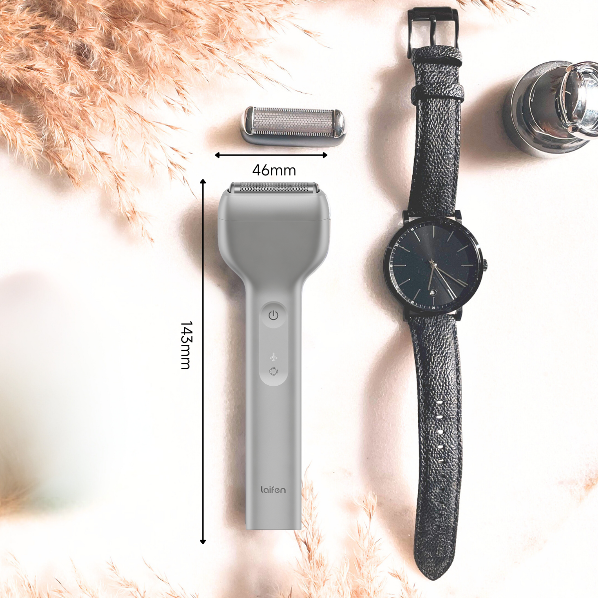 Un rasoio elettrico grigio, orologio e testina su una superficie bianca. Dimensioni indicate: 143 mm e 46 mm. Piante secche sullo sfondo.