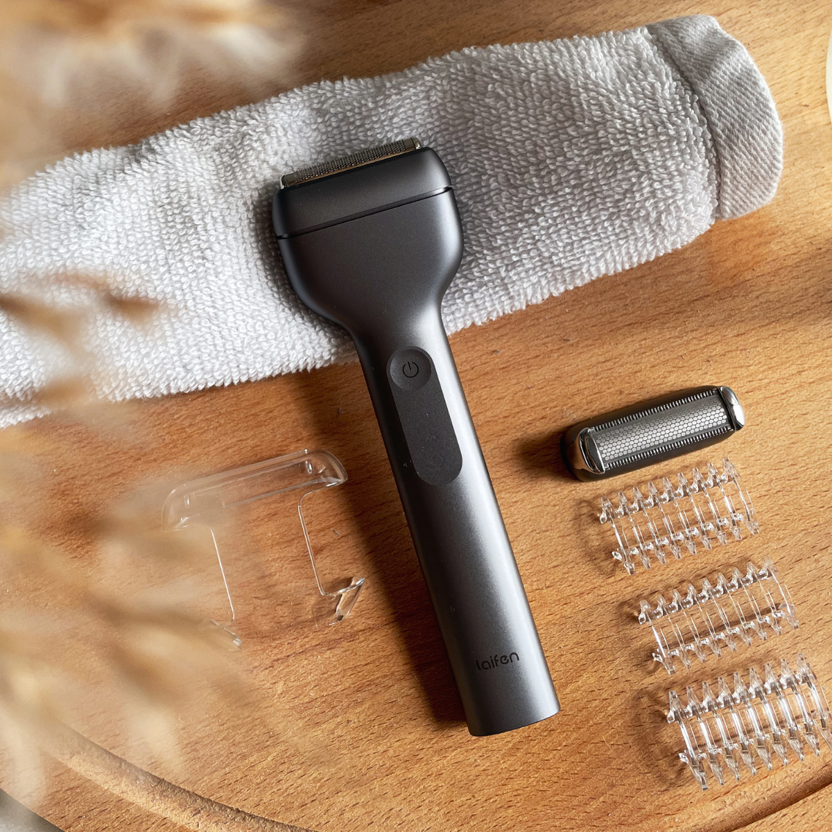 Een donkergrijze Laifen body shaver met accessoires op een houten oppervlak met een handdoek. Inclusief messen en doorzichtige plastic beschermkappen.