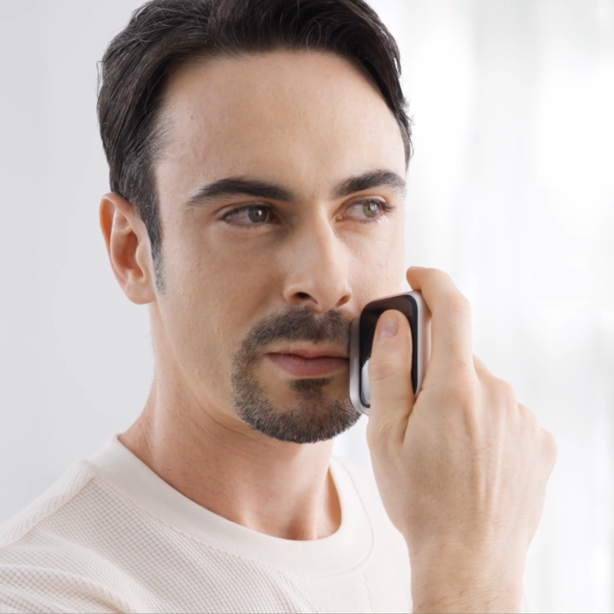 Un homme se rase le visage avec un appareil. Il porte un t-shirt blanc gaufré. Le fond est blanc.