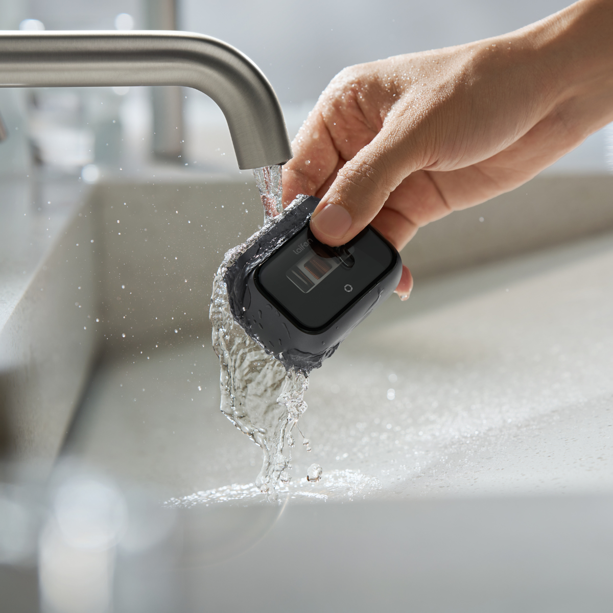Une main rince un appareil noir sous l'eau courante d'un robinet argenté. L'eau éclabousse l'évier blanc.