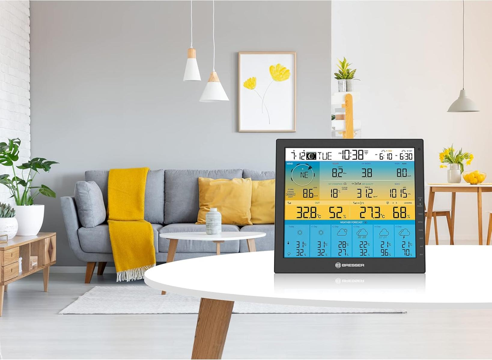 Ein Wohnzimmer mit Wetterstation auf einem weißen Tisch. Ein graues Sofa mit gelben Kissen und einer gelben Decke. Ein Gemälde an der Wand.