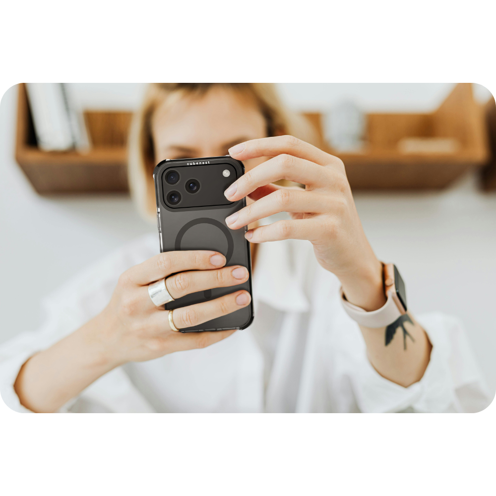 Eine Person hält ein schwarzes Telefon mit Hülle. Sie trägt ein weißes Hemd, Ringe und eine Uhr. Im Hintergrund befindet sich ein Holzregal.