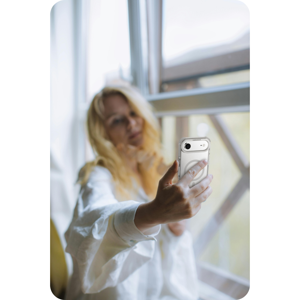 Eine Frau mit blonden Haaren macht ein Selfie mit einem Telefon in einer durchsichtigen Hülle. Sie trägt ein weißes Hemd und sitzt am Fenster.
